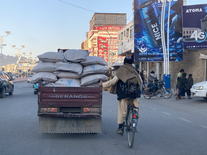 This image depicts a bustling street scene in Kandahar, Afghanistan, at what appears to be late afternoon or early evening.
In the foreground and midground, a reddish-brown flatbed truck, marked "BERANG" on its tailgate, is heavily loaded with numerous white sacks, many labeled "WHITE CRYSTAL RICE." The sacks are piled high, creating an unstable-looking load. To the right of the truck, a man wearing traditional attire, including a tan shawl, a dark vest over a light tunic, light-colored pants, and a dark cap, rides a black bicycle. He appears to be holding onto the side or rear of the moving truck with his right hand, with a dark backpack featuring a white grid design on his back. The truck's taillights are illuminated.
The paved road features multiple lanes, with a concrete divider on the left side where modern streetlights equipped with solar panels are visible. In the background on the left, other vehicles are present, and distant mountains rise under a clear, pale sky.
On the right side of the street, a commercial area is evident with multiple buildings and prominent billboards. A large billboard advertises "NICE" energy drink with a blue bottle design and Arabic script. Below it, another sign for "ATOMA 4G" is visible. Partially obscured signage for "KARIM BROTHERS" is also present on a building. Further right, a person on a motorcycle, wearing a face covering, is visible. Several pedestrians, including children, are walking near the roadside on the far right, where a white car is also partially visible. The overall environment suggests a busy urban thoroughfare with a mix of vehicle and pedestrian traffic.