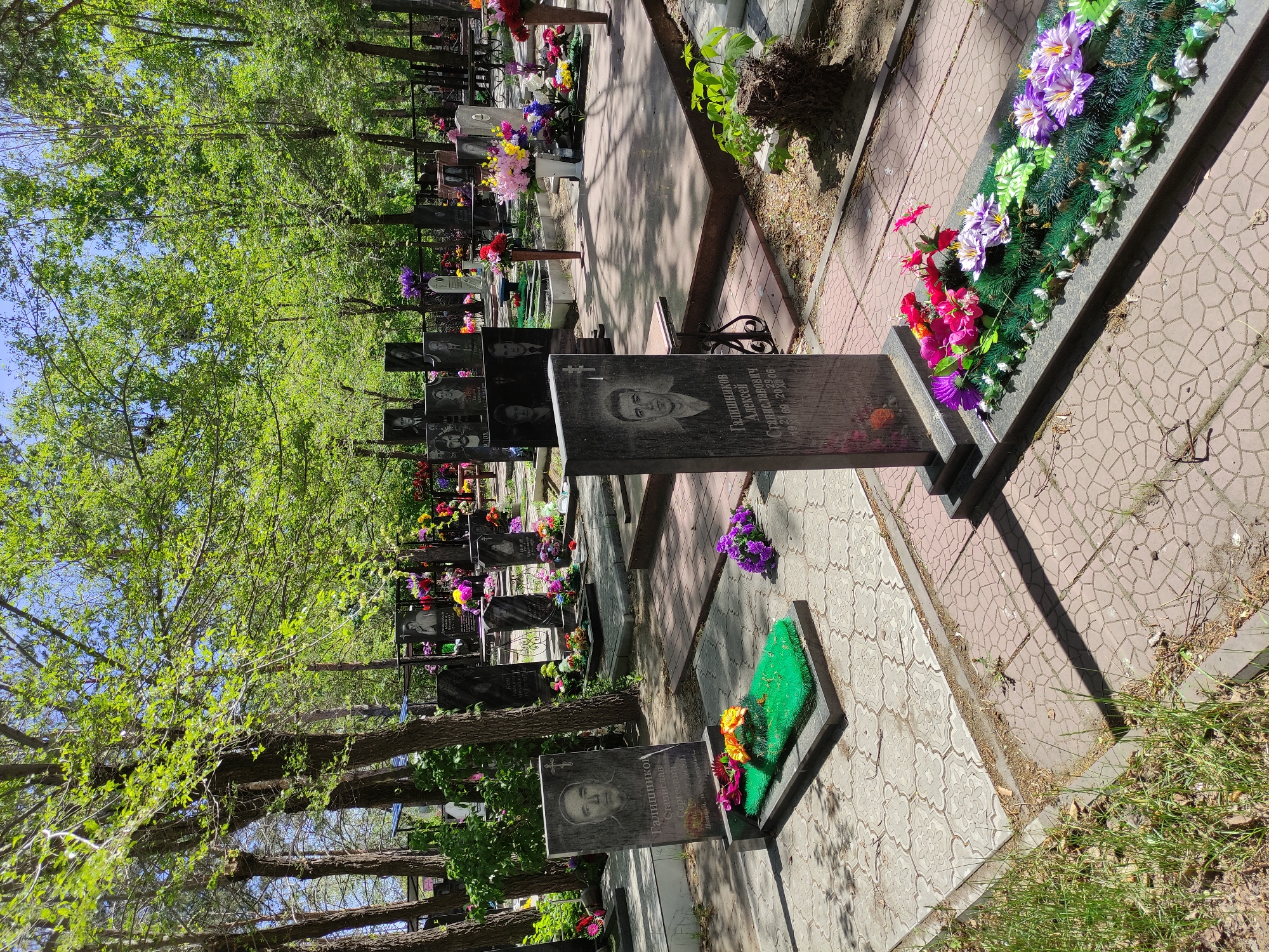 The image shows a cemetery in Pavlodar, Kazakhstan, with rows of tombstones lining a paved walkway. The tombstones are made of dark stone, some featuring photographs of the deceased. Artificial flowers adorn many of the graves. Trees with green foliage provide shade over the area, and some greenery grows along the edges of the walkway.
