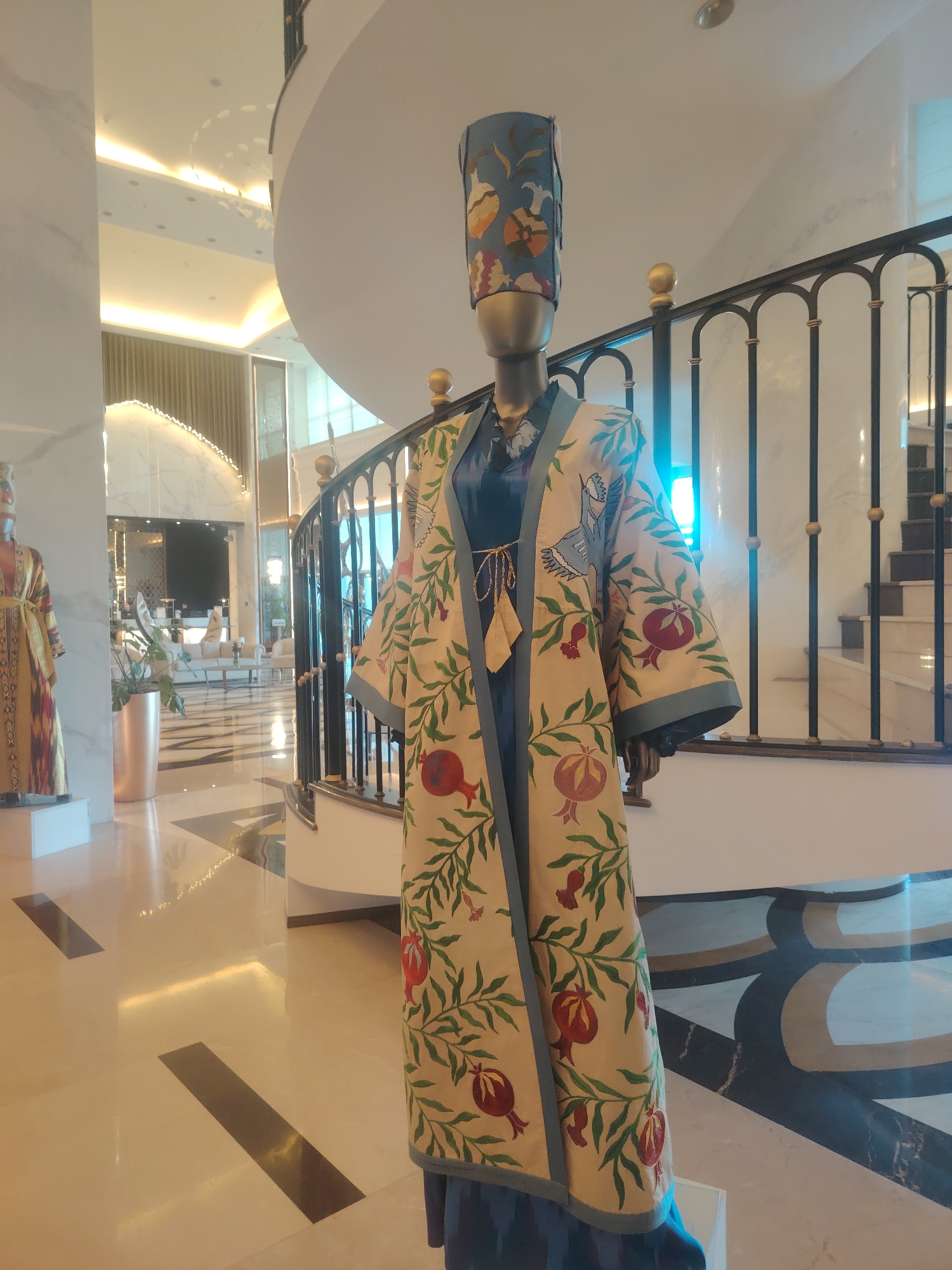 The image shows a mannequin wearing a traditional Uzbek robe with a tall, cylindrical hat. The robe is cream-colored and adorned with an intricate pattern of green leaves, red pomegranates, and blue birds. The mannequin stands on a polished marble floor in what appears to be a hotel lobby or a museum in Konigil, Uzbekistan. To the left, another mannequin in a similar traditional outfit is visible. A grand staircase with a black metal railing curves upwards in the background, and soft lighting illuminates the spacious interior, reflecting off the polished surfaces.