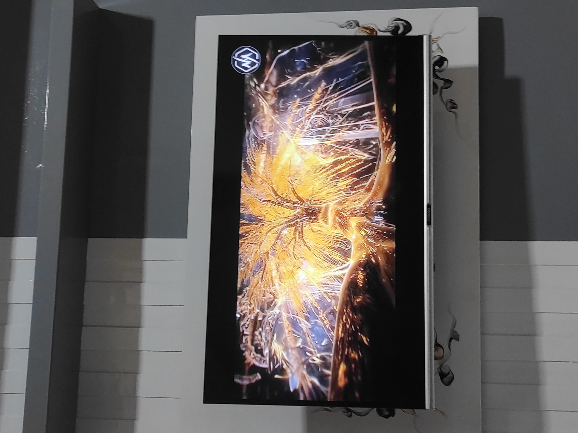 A television is mounted on a wall. The television screen displays a vibrant, golden-orange, fiery image with swirling blue and silver elements. A circular logo with the letters "NA" in white is visible in the upper right corner of the screen. The wall behind the television has a textured pattern of alternating light and dark gray vertical panels, with a darker gray band above. A white border frames the television, with a decorative, swirling black pattern extending onto the wall below the screen on the right side. The bottom edge of the television has a thin silver bezel with some visible text, possibly a brand name. The overall scene suggests an indoor setting, likely a living room or entertainment area, in Sumedang, Indonesia.