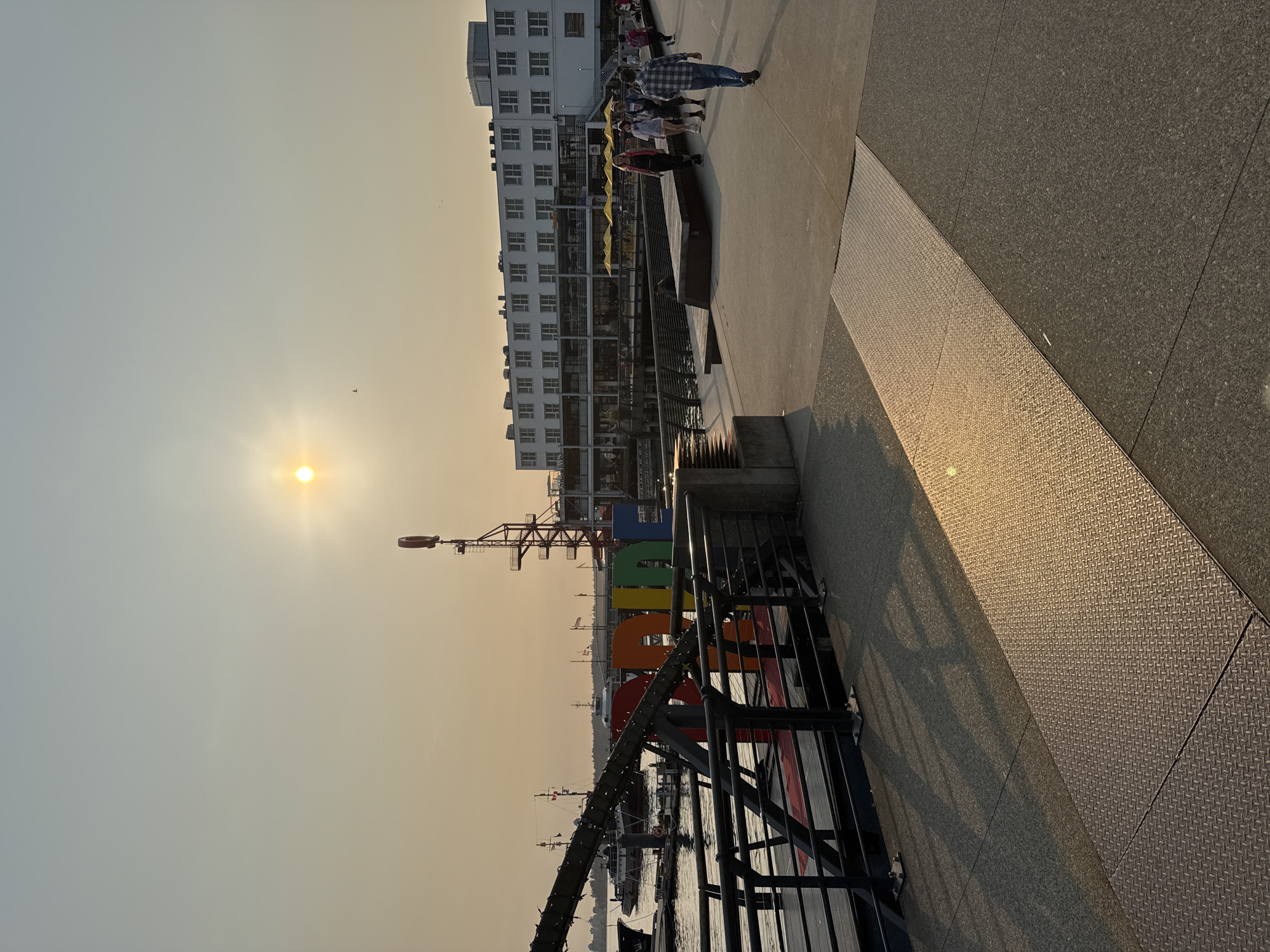 This image depicts a waterfront scene in North Vancouver, Canada, likely on a hazy or smoky day given the diffused sunlight. In the foreground, a paved walkway runs diagonally, with a section of diamond-plated metal flooring reflecting the golden light of the sun. To the left, a railing separates the walkway from a body of water where boats are docked. A large, colorful "PRIDE" sign made of individual, three-dimensional letters is visible on the left side of the frame, with some of the letters partially obscured by a metal structure.
In the mid-ground, a white multi-story building with many windows lines the waterfront. Several people are walking along the paved area in front of this building. The sun is positioned high in the sky, appearing as a bright, diffused orb due to atmospheric conditions. A tall, industrial-looking tower with a circular element at its top is also visible behind the "PRIDE" sign. The water appears calm, reflecting the sky and surrounding structures. The overall atmosphere is one of a public urban waterfront during daylight hours.