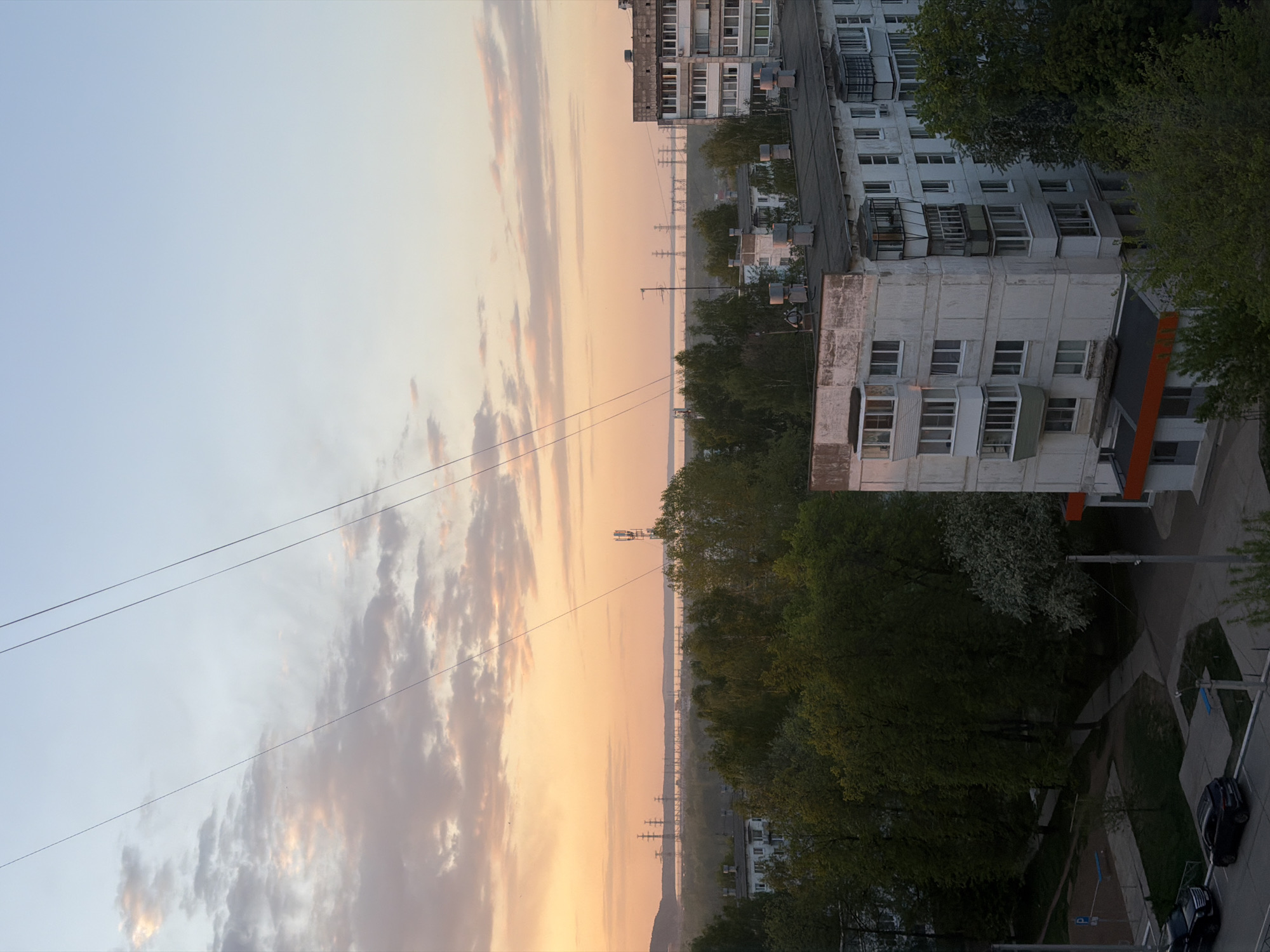 The image shows an urban scene during sunset in Chaikovsky, Russia. The view is from an elevated position, possibly a high-rise building. In the foreground, there are residential buildings with white walls and balconies. Green trees are interspersed among the buildings. The sky is visible in the background, displaying a colorful sunset with clouds and a soft orange glow. Power lines stretch across the sky. A river or large body of water is visible in the distance, bordered by trees. Power line support towers are spaced out over the river. There is a paved area with parking spots and cars visible in the lower right corner.