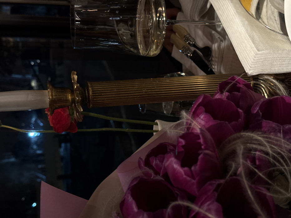 The image depicts a close-up, dimly lit indoor table setting in a restaurant or dining establishment in Yerevan, Armenia, at night. In the foreground left, a large bouquet of dark purple tulips, partially wrapped in light pink and beige translucent paper, contains white fibrous decorative material. Centrally, a fluted brass candelabra holds an unlit white taper candle. Adjacent to the candelabra, a small white vase contains two red flowers with slender green stems. To the right, a clear wine glass holds a pale yellow liquid, consistent with white wine. Below the wine glass, a silver knife and fork with ornate, light-colored handles rest on a folded white cloth napkin. A stack of white paper napkins is visible to the immediate right. The background is a dark, blurred view through a window, revealing out-of-focus blue and white streetlights and indistinct urban shapes. No individuals are visible.