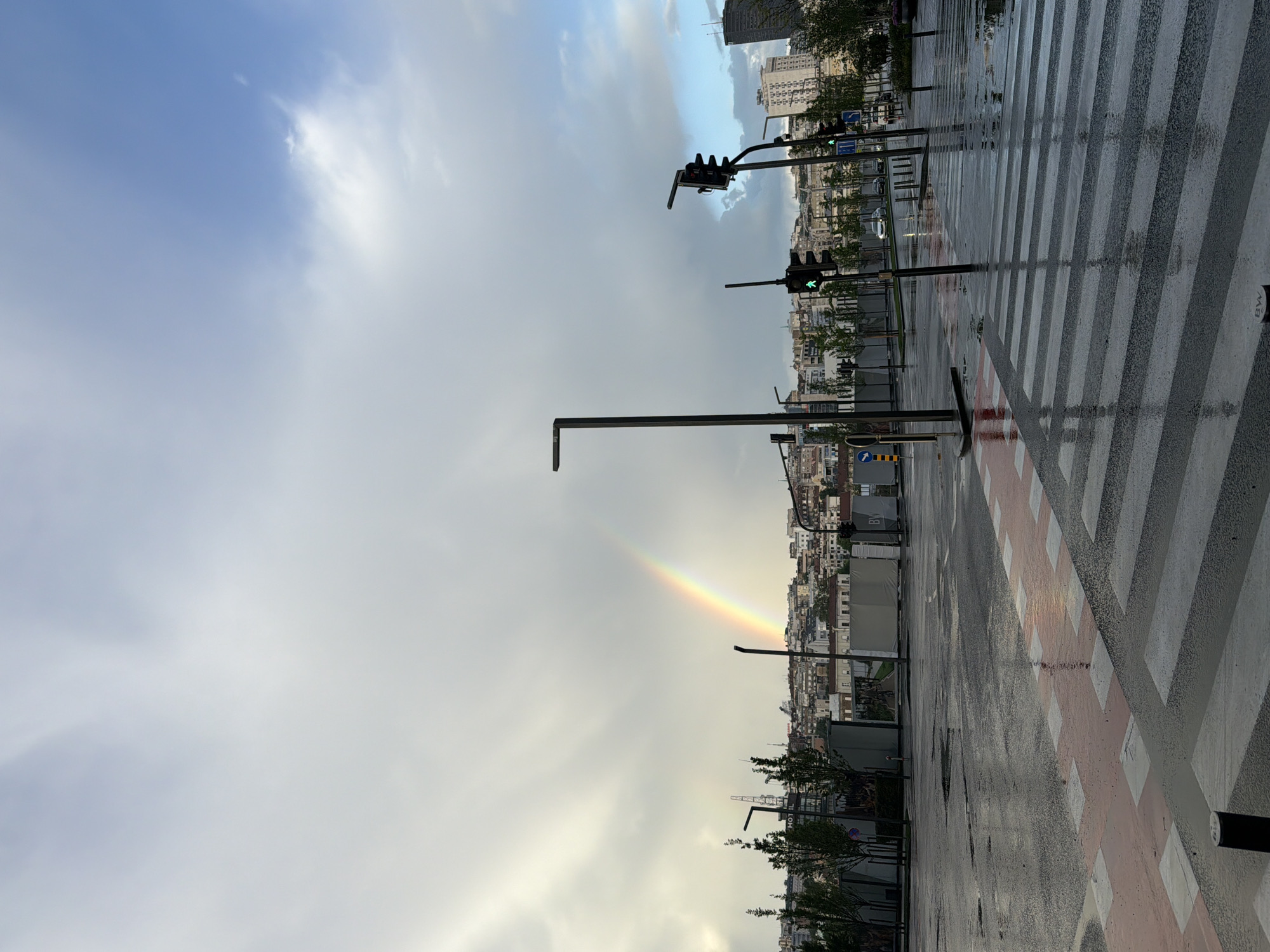 The image captures a street scene in Belgrade, Serbia, on a rainy day. A vivid rainbow arcs across the sky, emerging from the cityscape. The street is wet, with reflections on the pavement and pedestrian crossing. Traffic lights are visible with green lights. Buildings of varying heights and styles form the urban skyline. The sky is partly cloudy.