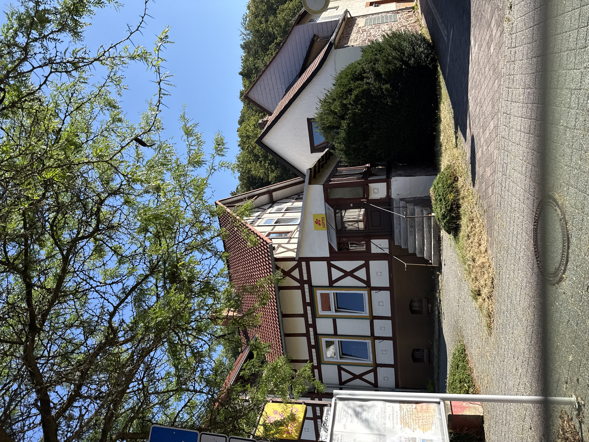 A sunlit exterior view of a half-timbered building with a red tiled roof in Bad Sachsa, Germany, is framed by the branches of a leafy tree. The building features a white facade with dark brown timber framing, and a brown base. Two windows with white and yellow trim are visible on the lower floor. Above them, on the first floor, are more windows. A small glass-enclosed entrance with a "LOTTO" sign is positioned to the right of the windows. To the right of the half-timbered section is a white building with a contrasting gray tiled roof. A dense green shrub sits in front of this section. The ground in front of the buildings is paved with cobblestones and gravel. A metal pole with signs is located to the left of the frame, and a street runs along the right side of the image. The background is filled with a dense, green forest. The sky is clear and blue.
