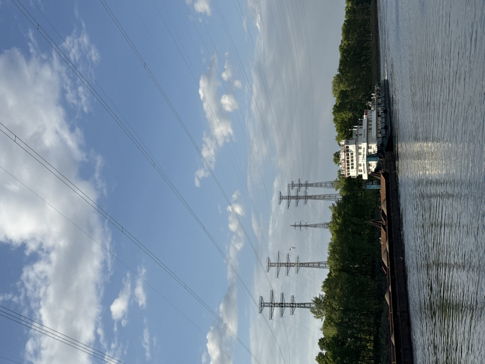 The image depicts a landscape with a river, a barge, power lines, and the sky. A barge is traveling on a river next to a tree-lined shore. Several power line towers are visible on the land, with power lines extending across the sky. The sky is blue with scattered clouds. The image is taken outdoors in daylight. The location is Chaikovsky, Russia.