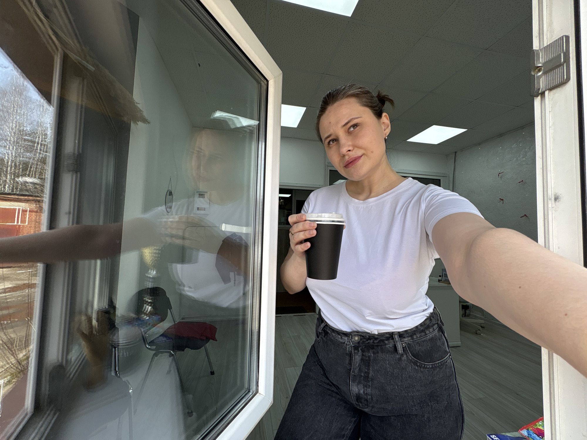 A woman is standing in an open doorway, holding a black coffee cup. She has her hair pulled back in a bun and is wearing a white T-shirt and black jeans. The reflection in the window shows a table and chair. Outside the window, there is a view of trees and a brick building. The interior of the room has a white ceiling and walls. The location is Chaikovsky, Russia, judging by possible clues in the reflection of the window.