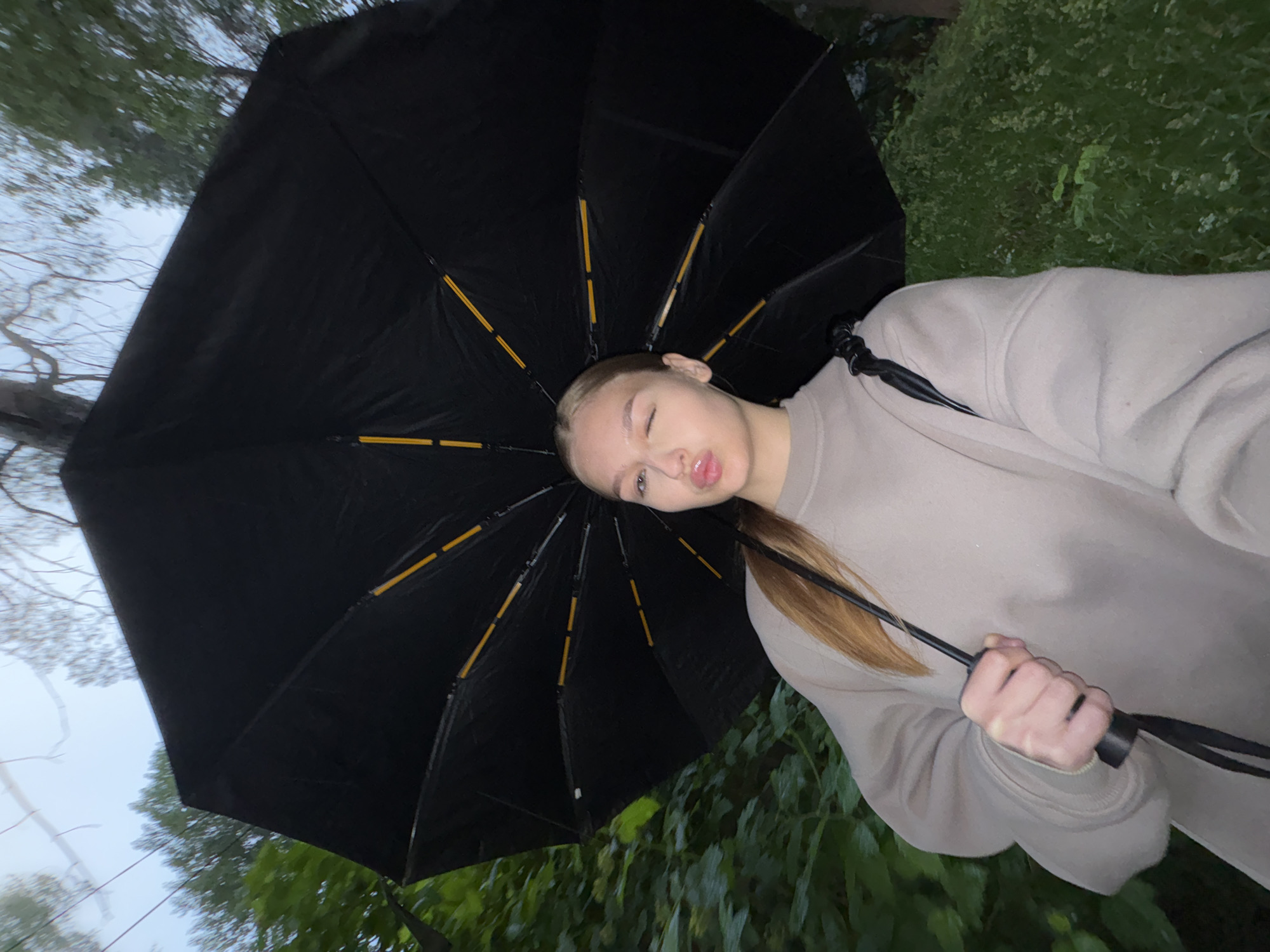 A young woman with blonde hair pulled back from her face is standing outdoors, holding a large black umbrella open above her. She is wearing a light brown sweatshirt and is winking at the camera with her lips pursed. The umbrella's spokes are visible, with yellow accents. Behind her, lush green foliage and trees are visible, suggesting a natural or park-like setting. The lighting is dim, and the sky appears overcast. This image was likely taken in Chaikovsky, Russia, given the provided context of the location.