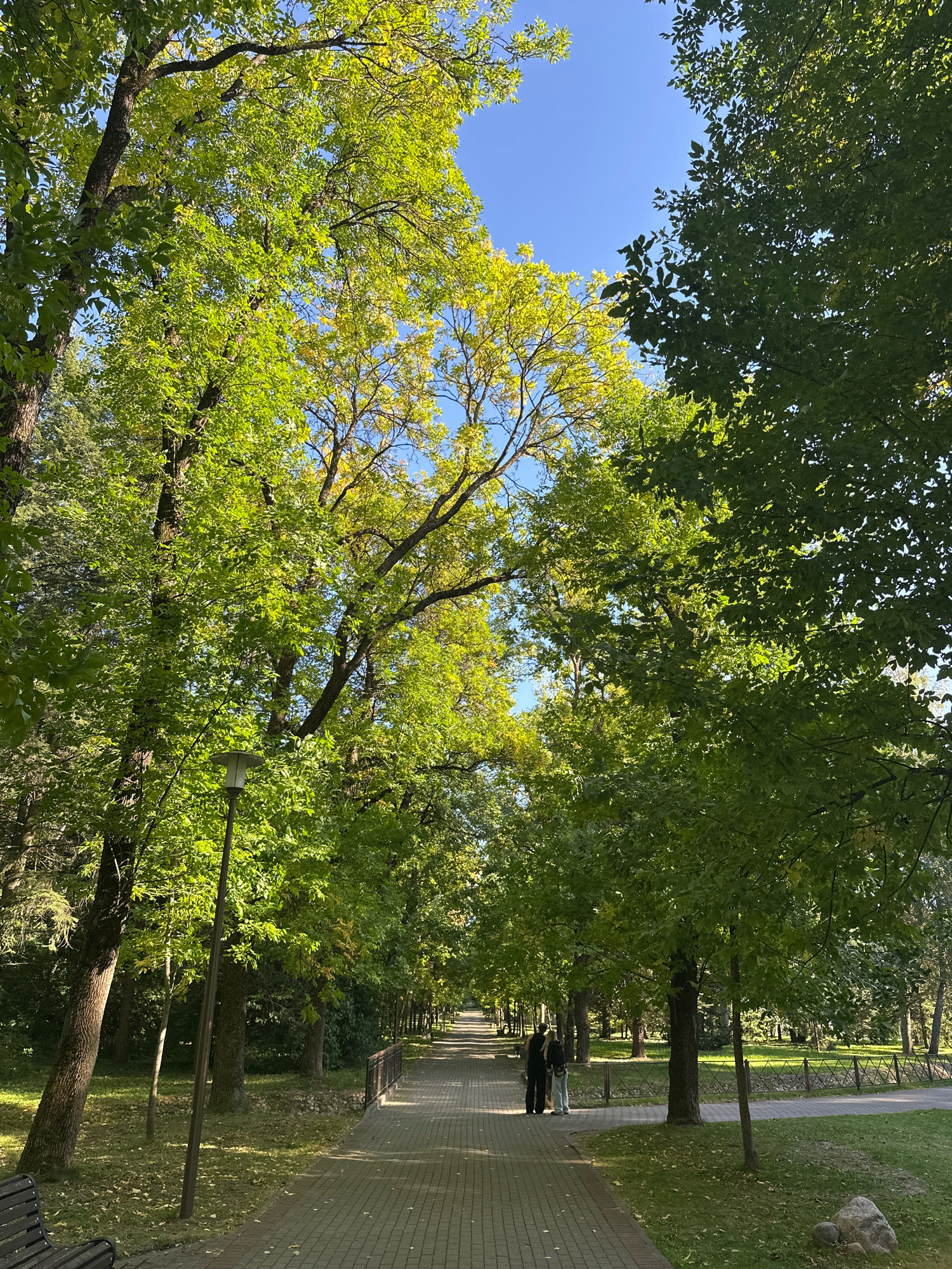 This is a photograph of a park with a paved walkway lined with trees. Two people are standing on the walkway, facing away from the camera. One person is wearing a dark shirt and pants, and the other is wearing a dark shirt and light-colored pants. A street lamp is visible to the left of the walkway. The trees have green and yellow leaves, suggesting it is autumn. The sky is blue. The scene is likely in the city of Minsk, Belarus, given the context.