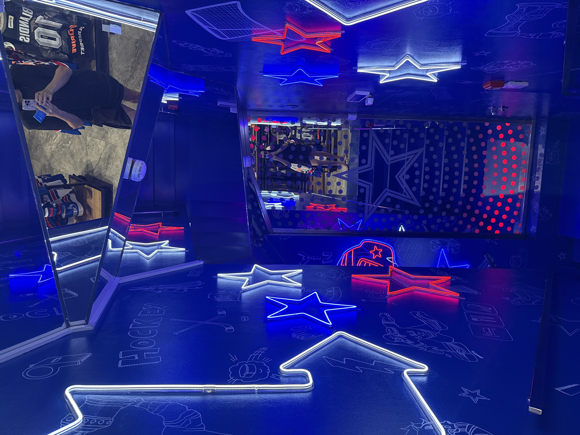 The image depicts an interior scene, likely a themed establishment, characterized by a vibrant blue wall adorned with white and neon-colored star and lightning bolt motifs. The word "HOCKEY" is also visible in a stylized font on the wall. In the background, a mirror reflects a person holding a smartphone, and racks of sports jerseys, possibly for sale. The environment appears to be a retail space or a recreational facility with a sports theme. The presence of neon lights suggests a modern and energetic ambiance. There is no specific information within the image to indicate its location in Saint Petersburg, Russia.