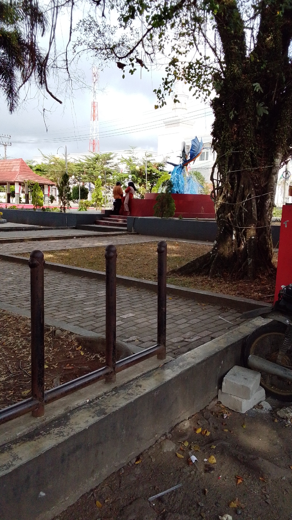 In Desa Karangbenda, Indonesia, a public outdoor space features a paved walkway with decorative metal posts in the foreground. Beyond the walkway, a tiered concrete structure supports a cluster of people in casual attire, one in an orange shirt and another in white. A large tree with moss-covered bark and exposed roots dominates the right side of the frame. In the background, a tall white and red communication tower is visible, along with buildings and dense greenery. A blue statue of a winged creature is positioned on a red pedestal. The ground is covered with dry leaves and dirt. A motorcycle wheel and exhaust pipe are visible in the lower right corner of the image. The sky is overcast.