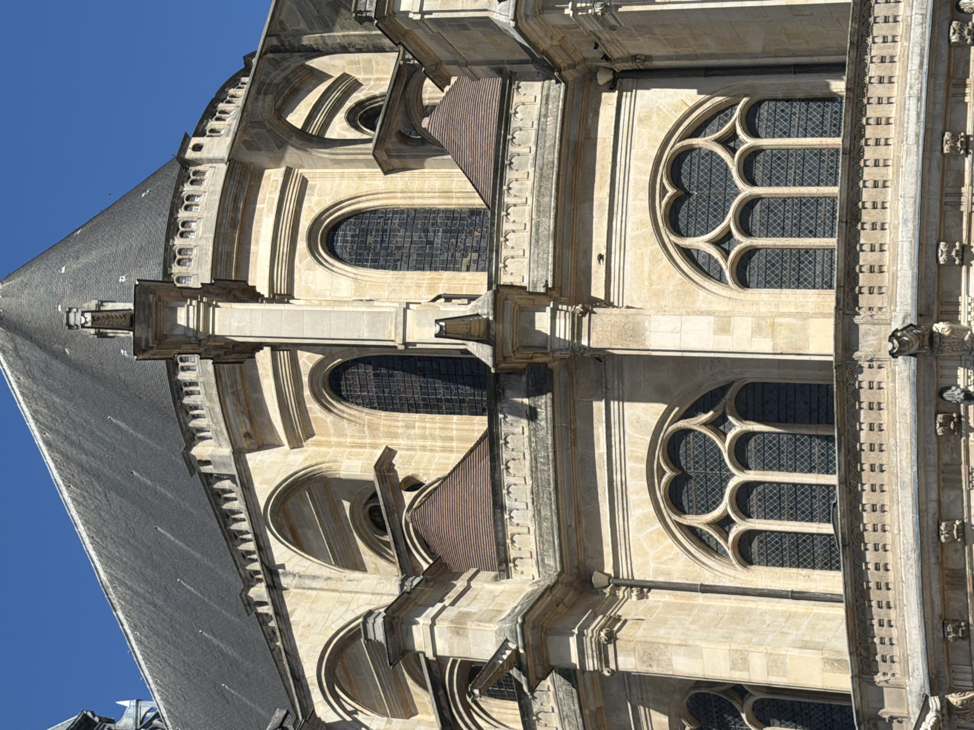 This is a close-up, oblique shot of the exterior of a Gothic-style building, likely a cathedral or church, with a dark gray slate roof and light-colored stone walls under a clear blue sky. The architecture features pointed arch windows, some with decorative tracery, and ornate stone carvings. A red-tiled roof section is visible lower down on the structure. The scene appears to be devoid of people.