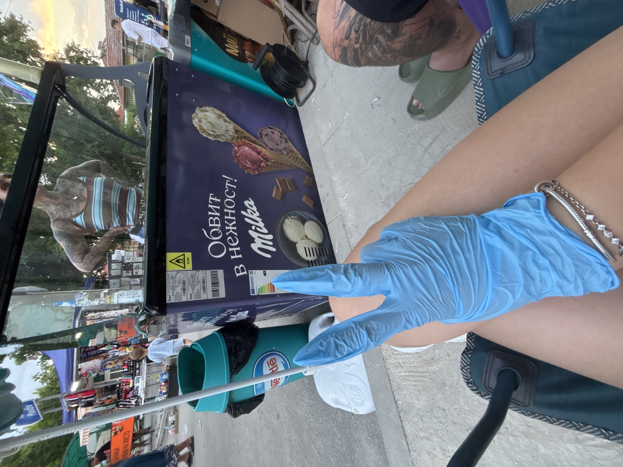 The image depicts an outdoor market scene in Kostievo, Bulgaria, observed from a low, seated perspective, likely in the late afternoon or early evening.
In the immediate foreground, a person's tanned right leg is visible, adorned with a multi-strand silver bracelet featuring small white stones. This person is wearing a light blue nitrile glove on their right hand, which is held up, displaying the index and middle fingers in a "V" or peace sign gesture. The individual appears to be seated in a dark green camping-style chair with a striped pattern. To the right, the lower leg and foot of another person are visible, wearing a dark green rubber slide sandal. This person's calf exhibits a prominent, intricate black and grey tattoo.
The midground is dominated by a dark blue upright ice cream freezer, prominently displaying the "Milka" brand name and the Cyrillic text "ОБВИТ В НЕЖНОСТ!" (Wrapped in tenderness!). Images of various ice cream cones and scoops, along with chocolate pieces, adorn the freezer. An energy efficiency label and a yellow warning triangle with "R290" are visible on its surface. To the left of the freezer stands a teal-colored cylindrical waste bin with the "Nestle" logo, containing a black trash bag. A white plastic base for a pole is situated next to the bin. To the right of the freezer, a black electrical cable reel rests on the paved ground.
The background reveals a bustling market environment. On the left, multiple market stalls with various merchandise, including clothing and trinkets, are set up under green and blue awnings. Several people are visible walking amongst these stalls, including a small child in a white outfit. A street sign displaying "Пловдив" (Plovdiv) and another partially visible sign reading "ОБЩИНА НА МЯСТО" (Municipality on Site) are present. On the right side of the background, a person with extensive tattoos on their arms and upper torso, wearing a brown and light blue striped tank top, is reflected in a glass panel above the ice cream freezer, appearing to stand behind a counter. Further back, a person in a white shirt and trousers stands near a building with a reddish-brown tiled roof. The ground throughout the visible area is paved.