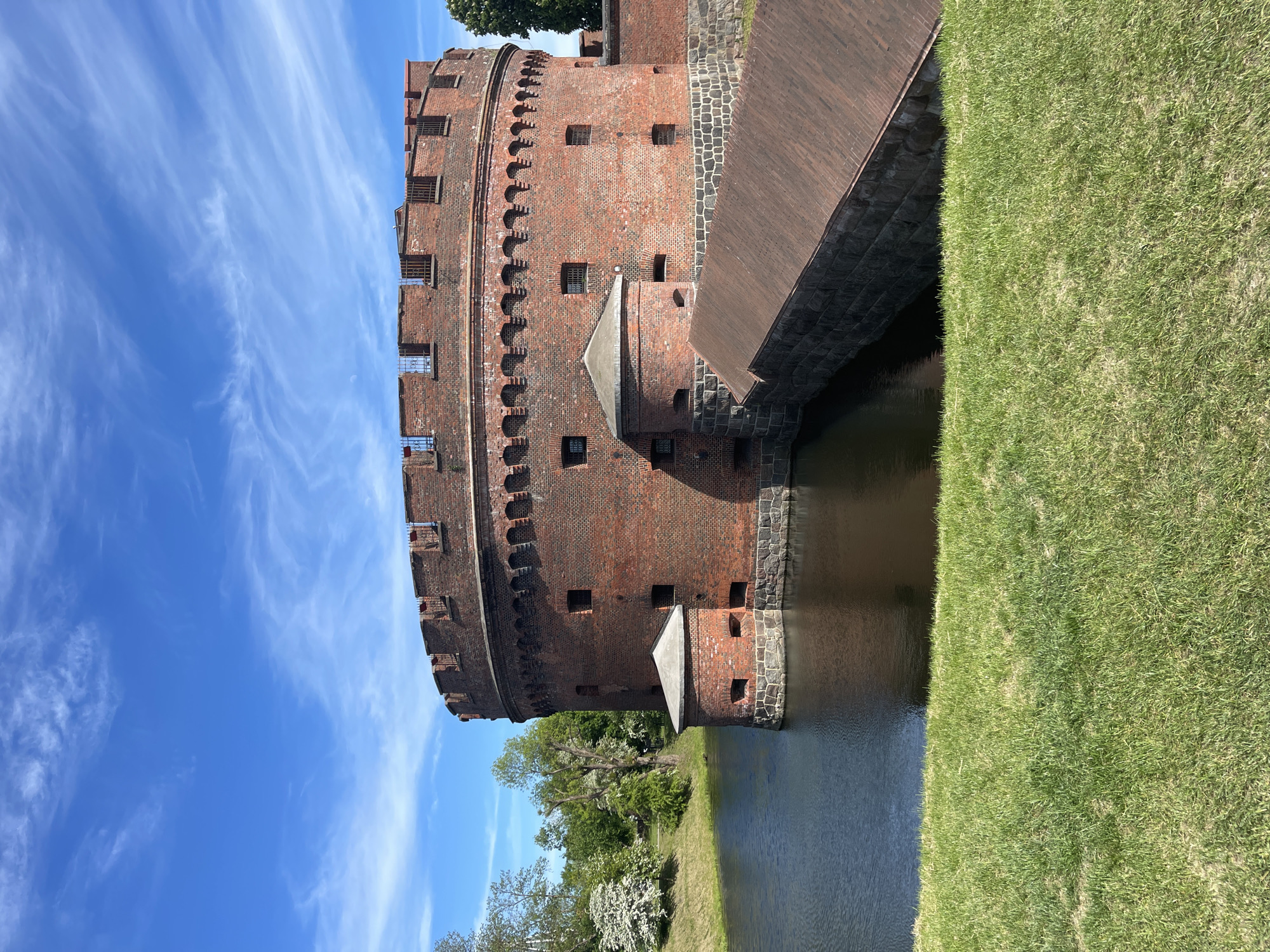 The image shows the Der Dona Turm, a red brick round tower in Kaliningrad, Russia. The tower is surrounded by water and a green grassy bank. A structure, likely a bridge or walkway, extends from the tower towards the grassy bank. The sky above the tower is blue with wispy white clouds. Trees with white blossoms can be seen in the lower portion of the image. The tower, water, bank, and sky occupy most of the scene.