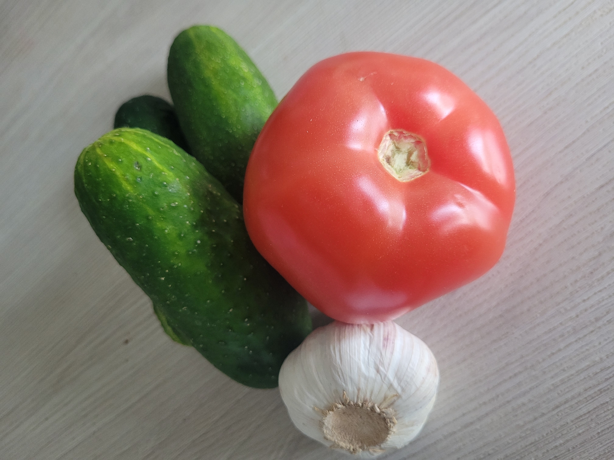 A close-up, overhead shot shows a collection of fresh produce arranged on a light-colored, wooden surface. In the foreground and slightly to the left, a whole head of garlic with a white, papery skin is visible. To the right of the garlic is a bright red, ripe tomato. Behind the garlic and tomato, three green cucumbers are nestled together, with their bumpy skins clearly discernible. The wooden surface appears to have a subtle grain pattern. The scene is well-lit, highlighting the textures and colors of the vegetables. The image is an indoor still life, with no indication of its specific location in Jūrmala, Latvia.