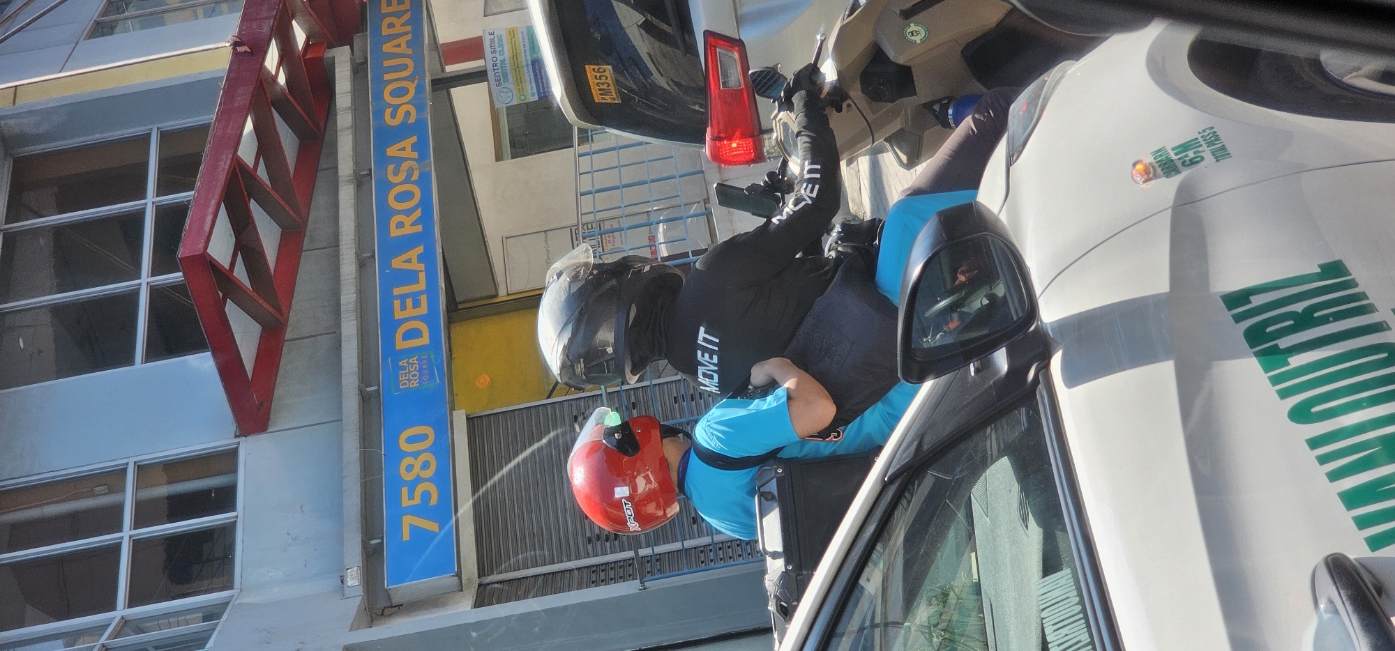 The image captures a scene on Dela Rosa Square in Makati City, Philippines, indicated by a prominent blue signage bearing "7580 DELA ROSA SQUARE". Two individuals are on a motorcycle, viewed from the side and slightly from the rear. The person in front wears a red helmet with "X-POT" branding and a bright blue t-shirt. They have a black bag slung over their shoulder. The person behind them wears a black helmet and a black hooded jacket with "MOVE IT" printed in white on the back and sleeve. They appear to be holding a mobile phone.
The motorcycle is positioned in front of a white car, with its rear visible. The car has a license plate reading "M356" and also features green lettering on its side, partially obscured but reading "MANIOUTRIZ" and "GARBAGE 6:M TOTAL PASS 5".
The background shows a building with large glass windows and a red awning structure. The overall environment suggests an urban setting, likely a street or parking area, under daylight conditions, with clear skies. The scene appears to be captured during daytime.