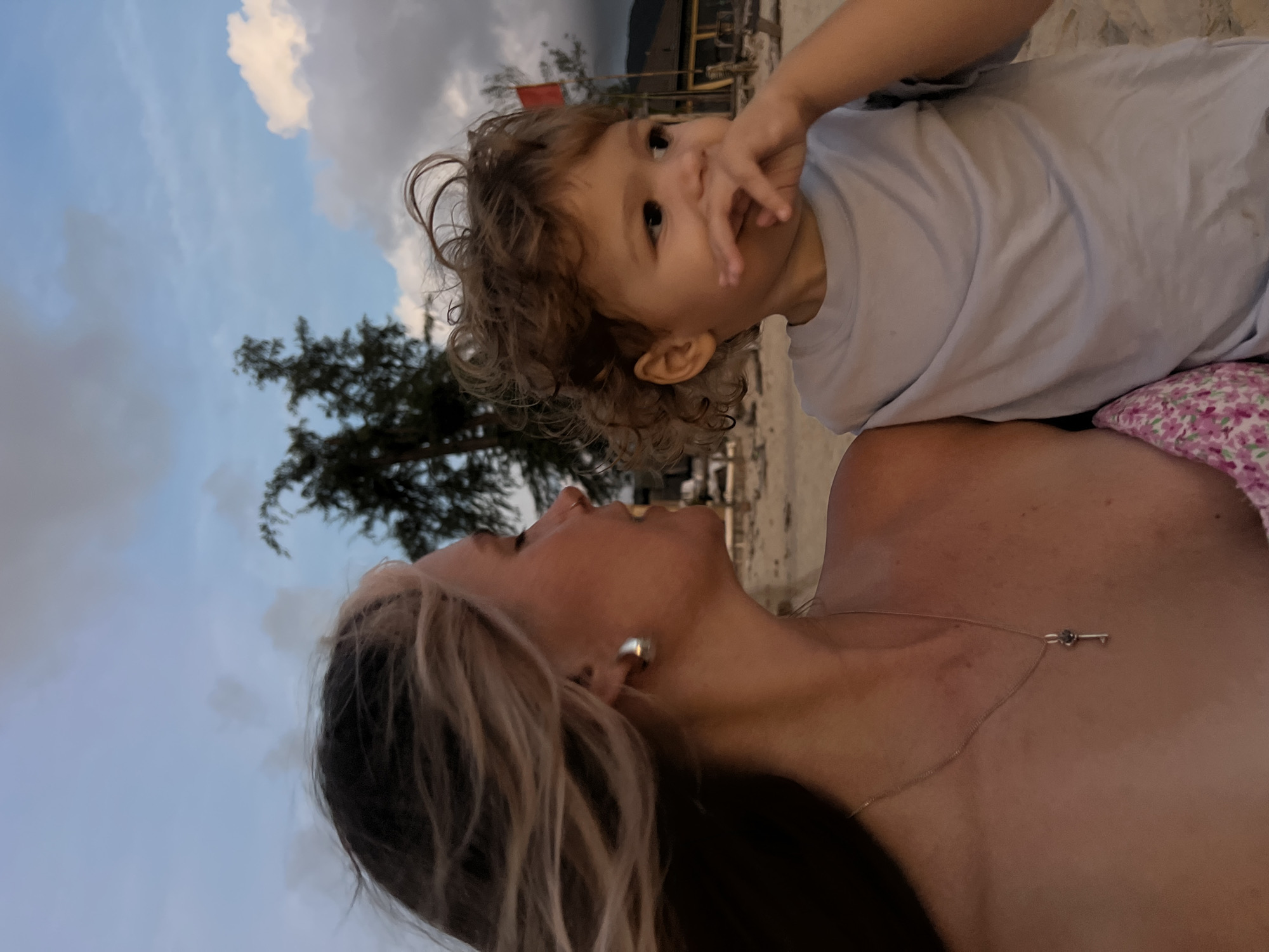 The image shows a woman and a young child on a beach, with the sky in the background. The woman is on the bottom, with her head tilted back and her eyes closed. She has blonde hair and is wearing a necklace with a key pendant. The child is on top of her, looking towards the camera with their hand near their mouth. The child has curly brown hair and is wearing a light blue t-shirt. The scene appears to be at dusk, with a cloudy sky and a tree visible in the background. There is also some blurry structures in the far background, possibly beach huts or buildings. The overall mood seems to be intimate and loving, with the two subjects looking at each other.