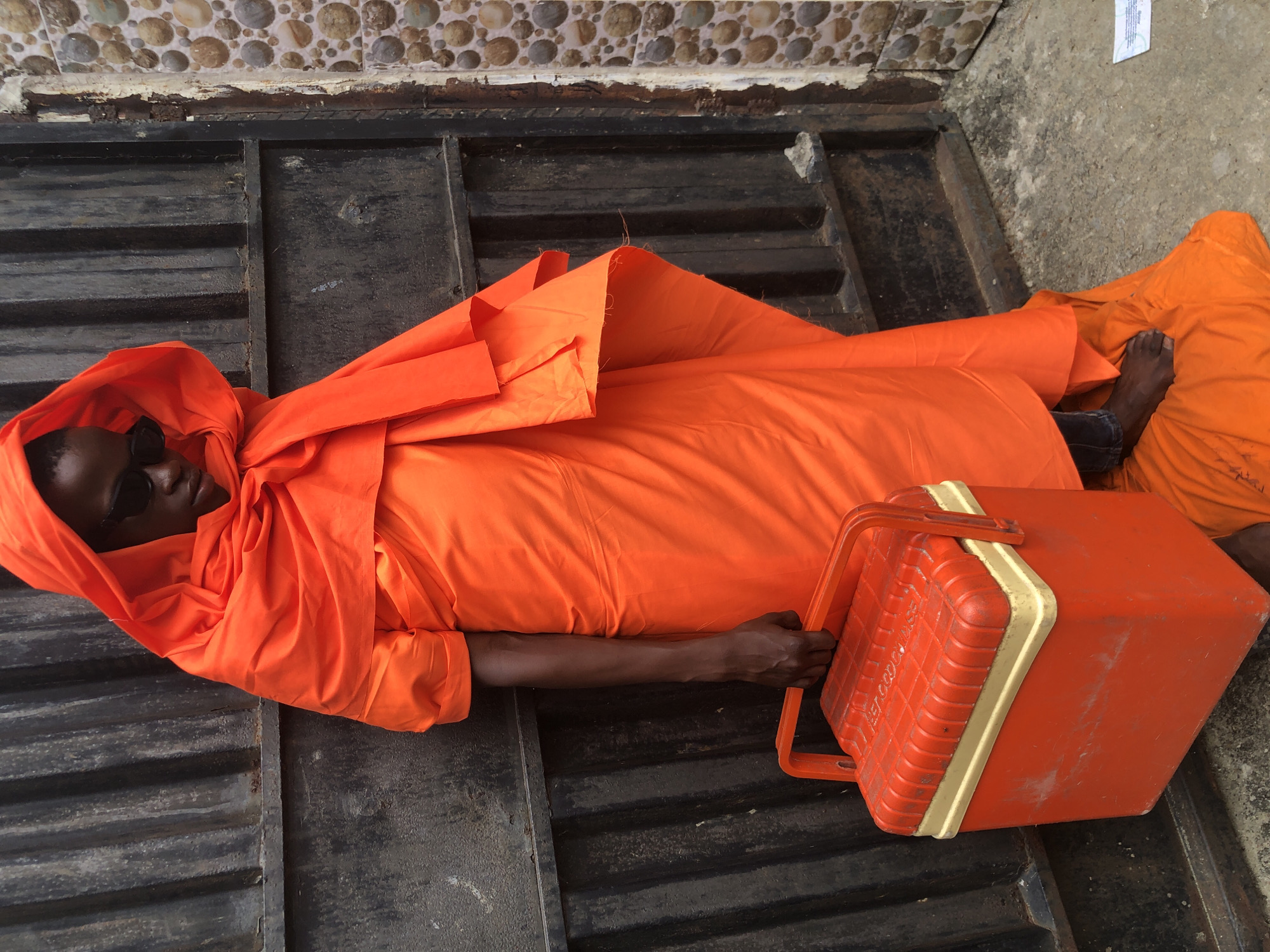 A person stands in front of a black metal gate in Ado, Nigeria, wearing a bright orange robe that covers their head and body, with sunglasses on. They are holding a red and yellow cooler box in their right hand. The person's bare left foot is visible on the ground, peeking out from under the robe. To the right of the gate, a wall is decorated with a pattern of round, multicolored stones. The ground is a concrete surface.