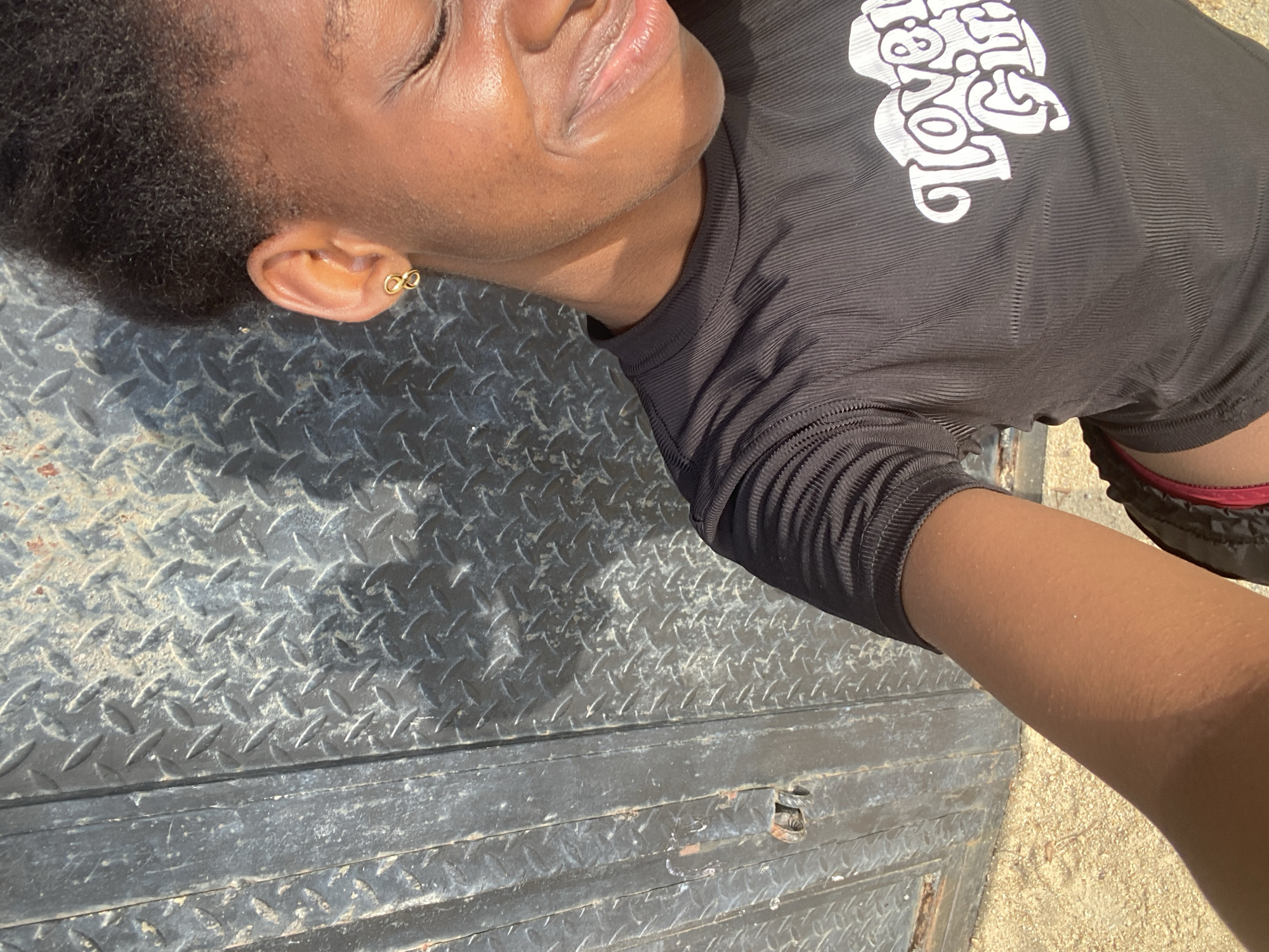 A close-up, angled shot shows a dark-skinned person lying on a metal surface. The person wears a black, ribbed long-sleeved shirt with white text on the upper right chest. A small, gold-colored earring is visible in the person's left ear. The metal surface beneath the person has a raised diamond plate pattern and appears weathered. The person's right arm extends into the frame. This image appears to be in Iranla, Nigeria.