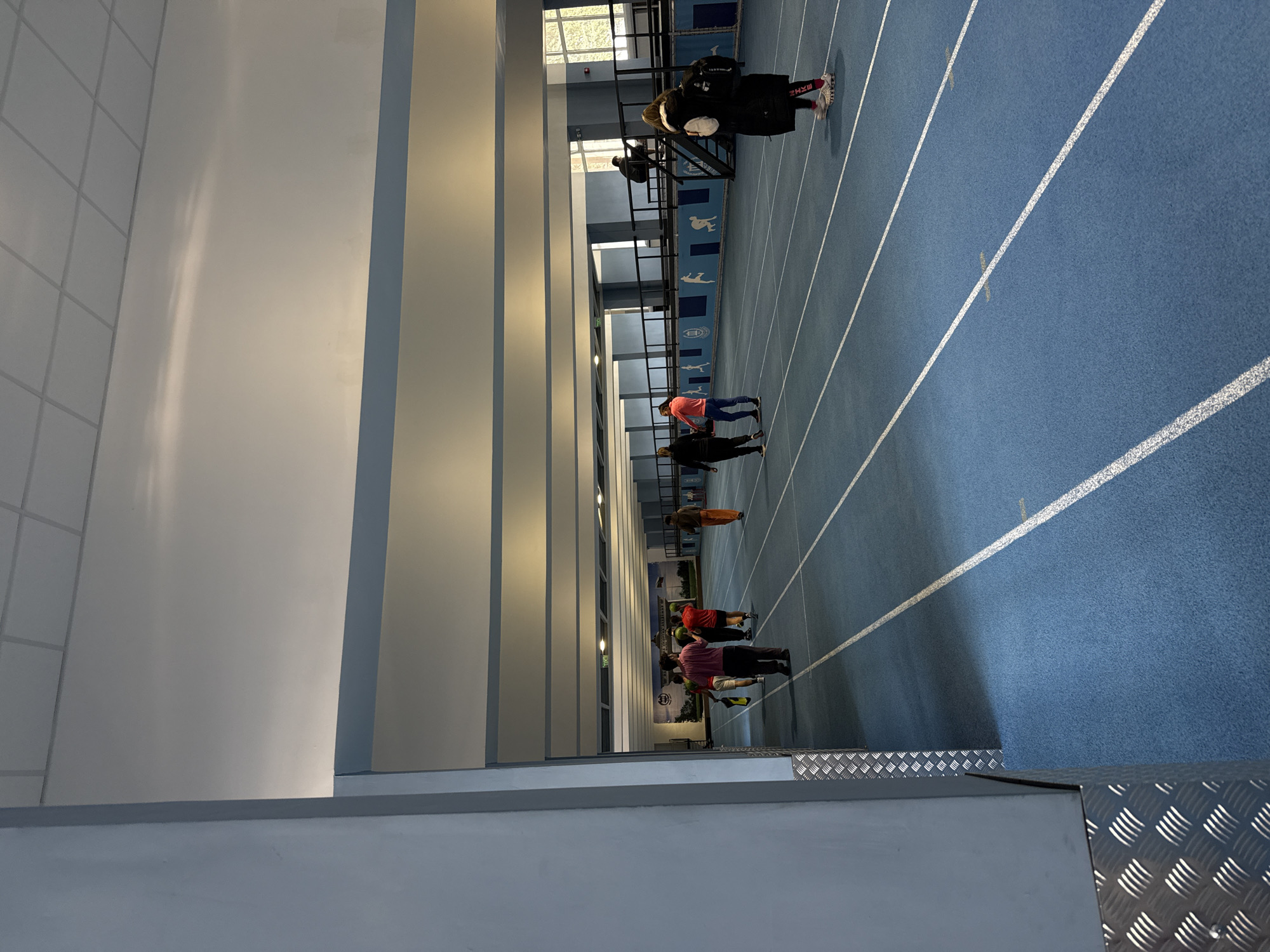 The image depicts the interior of an indoor athletics track facility, identified by a visible banner as the National Sports Academy "Vasil Levski" in Sofia, Bulgaria. The primary subject is a blue synthetic running track with white lane markings and intermittent yellow indicator lines.
Approximately nine individuals are present within the facility. On the left side of the track, a group of five individuals, primarily dressed in athletic attire, are walking from left to right. This group includes a person in a pink t-shirt, a person in a red t-shirt holding a green spherical object, a person in orange trousers, and a person in a green t-shirt holding a yellow rectangular object.
On the right side of the track, a female individual, dressed in a black long-sleeved puffer jacket, dark leggings with "NIKE" branding in pink/red lettering, and white sneakers, stands facing away from the camera, carrying a black backpack. Further to the right, an individual in dark clothing is crouched on the steps of a dark metal staircase that ascends to an elevated platform or spectator area. Two other individuals are partially visible further down the track on the right.
The environment includes light blue structural pillars in the immediate left foreground, featuring silver diamond plate metal cladding at their base. The walls are light-colored, with a continuous horizontal light blue band running above the track level. A white suspended ceiling with grid panels and integrated linear lighting fixtures covers the overhead space. Natural light enters the facility through large windows on the far right wall, revealing outdoor foliage. A large banner displaying the academy's name in Cyrillic, along with athletic silhouette graphics, is affixed to the far left wall. Dark metal railings define the elevated platform on the right side of the track.