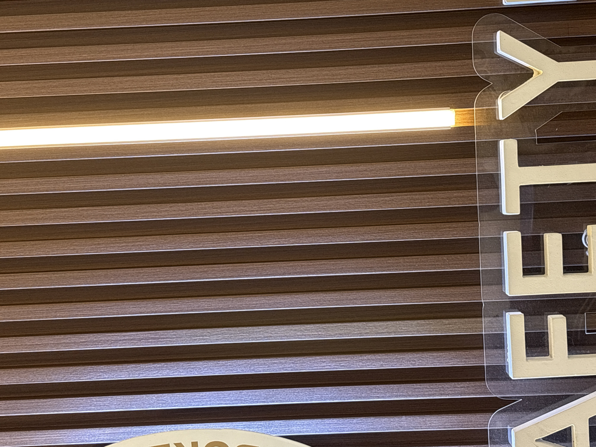 The image presents an interior scene, likely a commercial or public space, featuring a wall covered in horizontal, dark brown wooden slats. A bright, white LED light strip runs horizontally across the upper middle section of the wall, casting a clear glow and highlighting the textured grain of the slats.
On the right side of the image, a vertical sign is prominently displayed. It consists of large, white, three-dimensional letters mounted on a clear acrylic panel, creating a slight stand-off from the slatted wall. The visible letters of this sign, read from top to bottom, are "Y", "T", and "E". Below the "E", the top curve of another letter, possibly an "O" or "C", is partially visible.
In the bottom left corner of the image, a portion of a separate sign is visible. This sign appears to be light-colored with dark lettering, and the visible text consists of the letters "ON".
No people are present, and no specific activity or event is taking place; the image captures a static view of the architectural elements and signage within the space. The artificial lighting suggests an indoor setting where the time of day is not apparent.