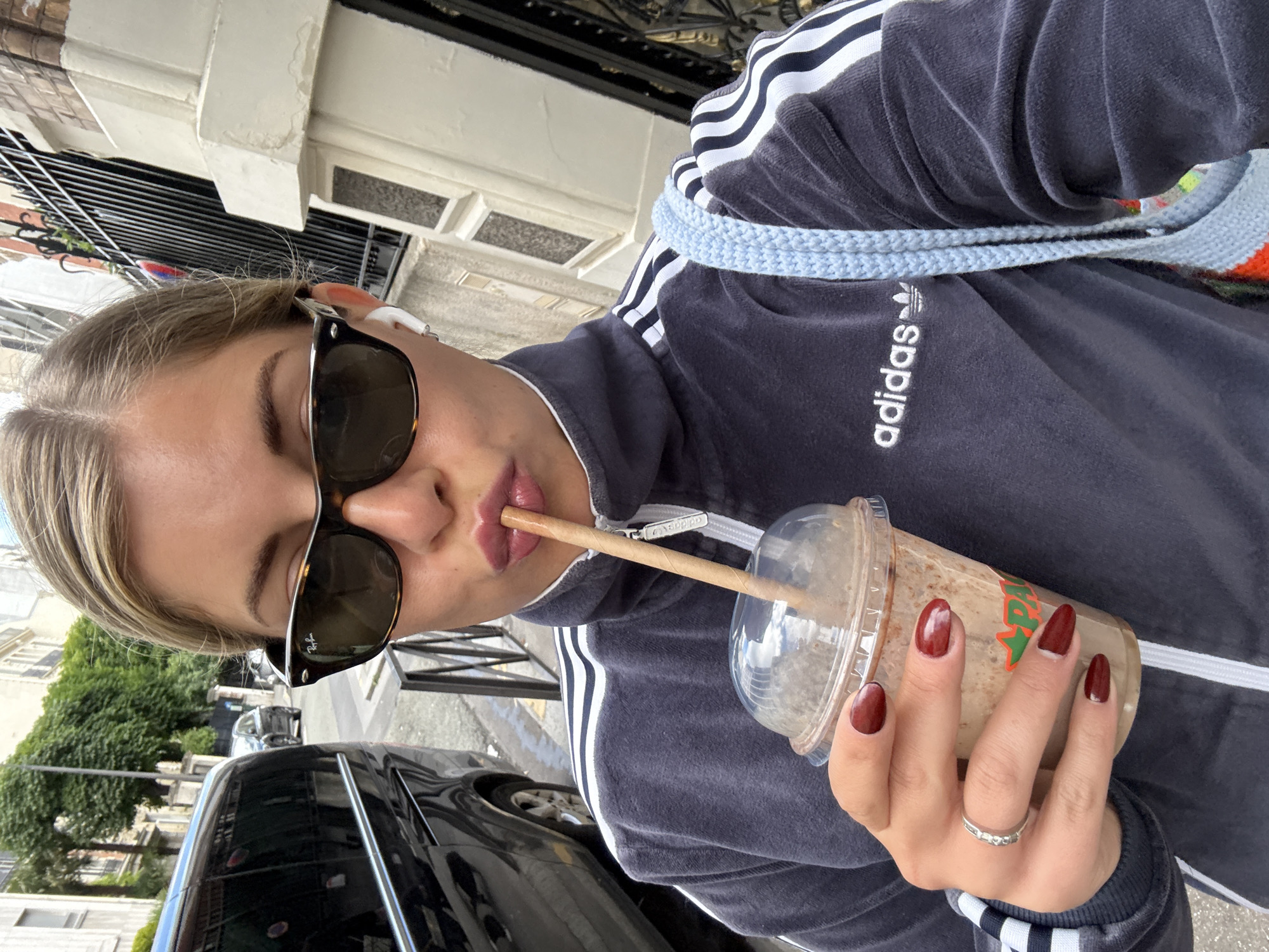 A young, fair-skinned woman, likely in her twenties, is taking a selfie while outside, potentially on a street in Paris, France. She's wearing sunglasses, an Adidas tracksuit, and is drinking from a clear plastic cup with a straw. Her nails are painted in a dark red color, and she has a ring on her finger. A black car is partially visible in the background, along with buildings and a wrought-iron fence, characteristic of Parisian architecture. The scene appears to be well-lit, suggesting it's daytime.
