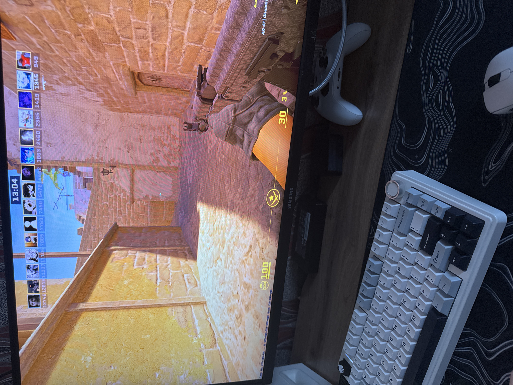 The image shows a close-up view of a computer monitor displaying the first-person perspective of a Counter-Strike: Global Offensive (CS:GO) game. The player is holding an AK-47 rifle, and the in-game heads-up display (HUD) is visible, including the player's health, ammunition count, and team scores. To the right of the monitor are various gaming peripherals: a white Xbox controller, a white computer mouse, and a custom mechanical keyboard with white and black keycaps. The peripherals are arranged on a dark-colored desk surface with a topographic pattern. It is not possible to determine the location in Perm, Russia from this image.