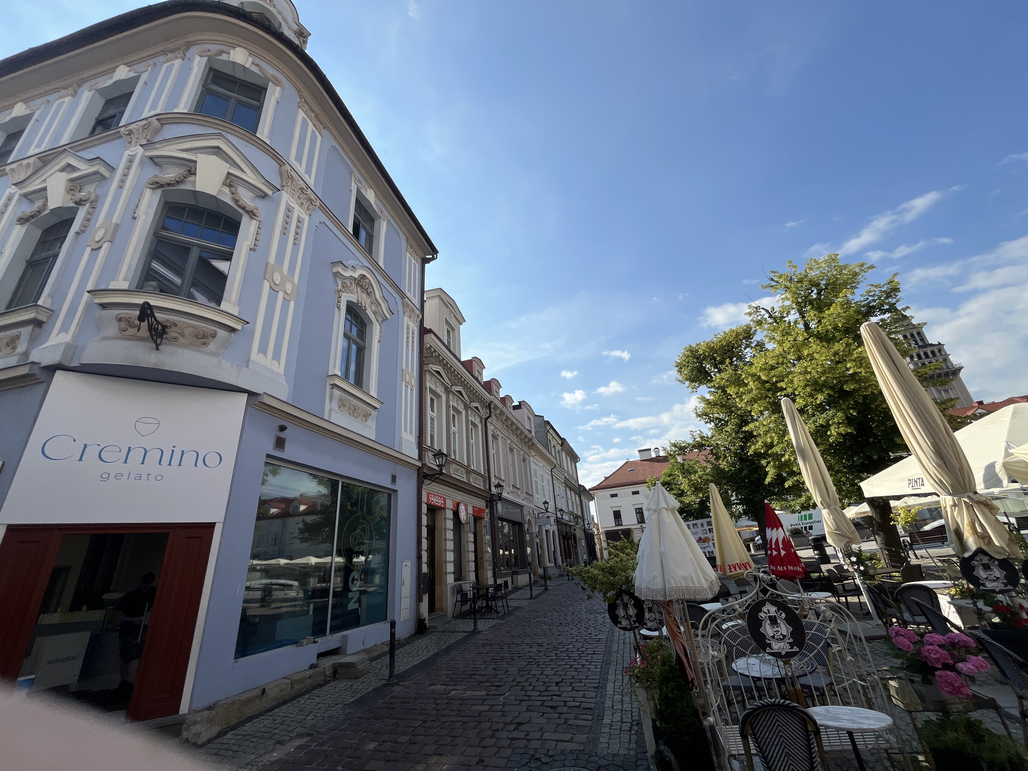 The image shows a street scene in a European city. On the left is a light blue building with ornate white trim and arched windows. A gelato shop called "Cremino" is visible on the ground floor, with its entrance and a large display window. The street is paved with cobblestones. To the right of the gelato shop, the street continues, lined with other buildings and outdoor seating areas for cafes. Several closed umbrellas and tables are set up outside. A large green tree is on the right side of the image, and a tall tower or spire is visible behind it. The sky is blue with scattered white clouds.