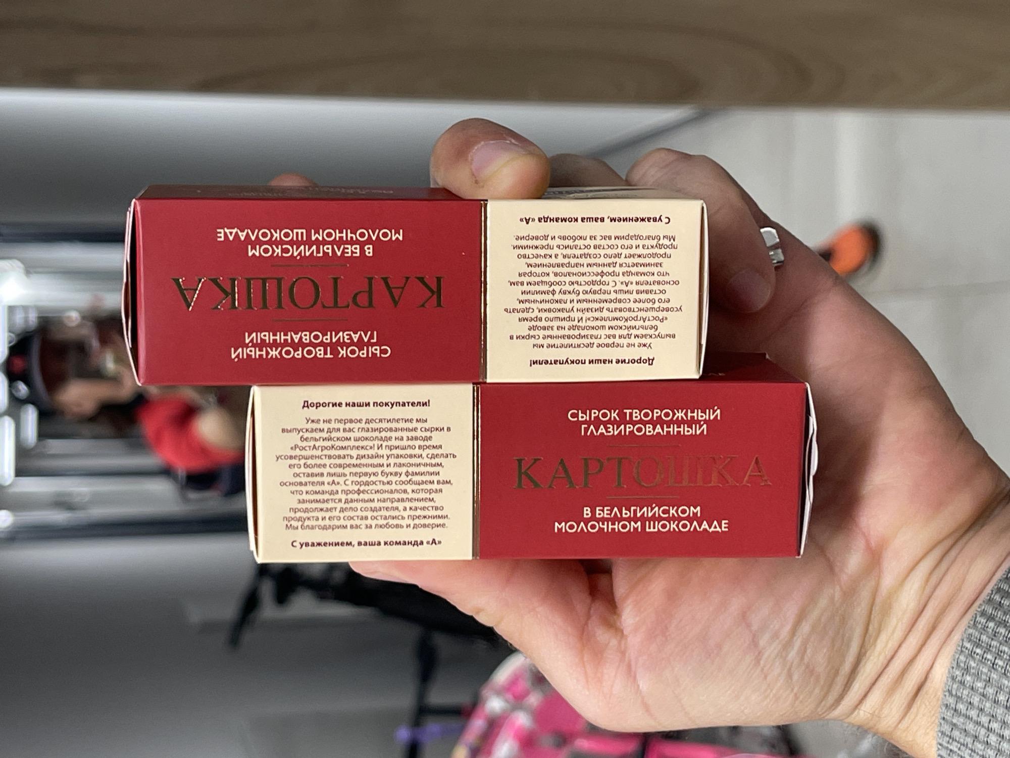 Here is a matter-of-fact description of the scene:
The image shows two red boxes of "Kartoshka" glazed curd cheese in Belgian milk chocolate. The boxes are being held by a hand with a ring on the ring finger. The text on the boxes is in Russian and describes the product and company. The background is slightly blurred, showing a glimpse of an interior space with a white wall and other indistinct objects.