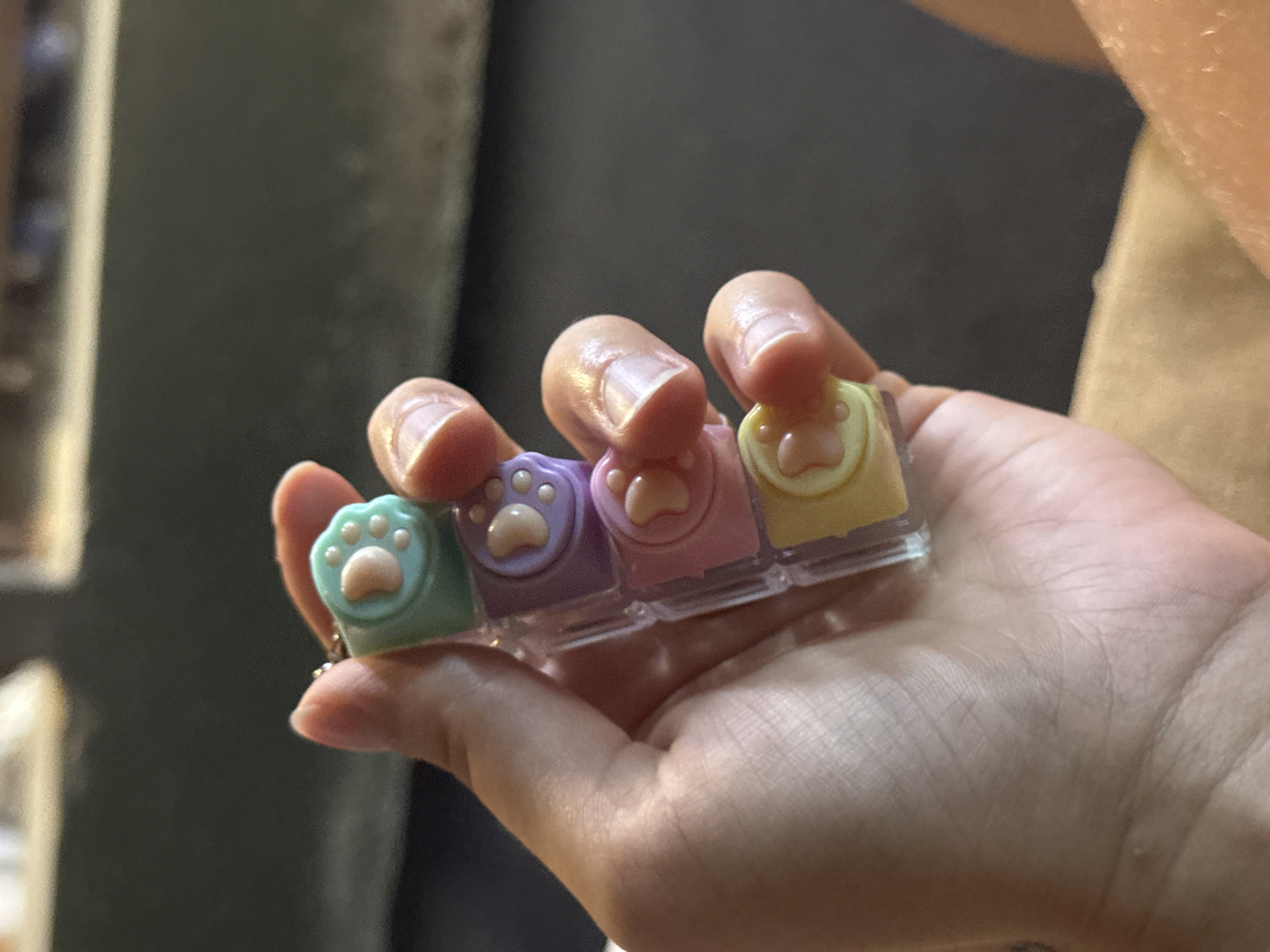 A hand is holding a small, clear plastic object containing four colorful, paw-shaped charms. The charms are stacked vertically and are colored light green, light purple, light pink, and yellow from top to bottom. Each charm features a pink paw pad and several small white dots representing toes. The background is out of focus and appears to be a dark, neutral color. This image was taken in Aksu, Turkey.
