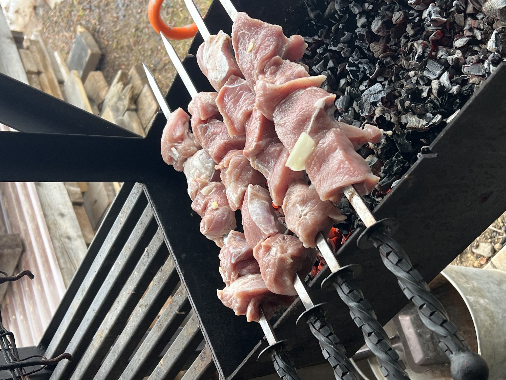 The image shows metal skewers loaded with raw meat chunks, likely pork or beef, resting over a charcoal grill. The coals beneath the meat are hot and glowing. The skewers have twisted, dark-colored handles. The grill is a black metal construction. The location is unknown.