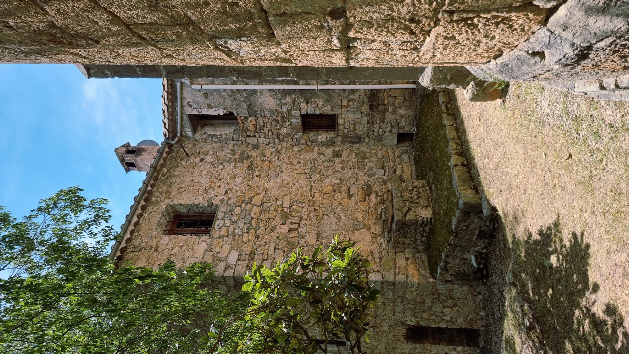 This image depicts a view of a rustic, stone-built structure with a tiled roof under a clear blue sky, likely in the town of Sales de Llierca, Spain. The foreground is a dry, grassy area with scattered pebbles and a prominent shadow cast by a tree on the left. The main structure, built from irregular-shaped stones, features a small window with a metal grate and a brick chimney protruding from the roof. Adjacent to this building, another stone wall, with much larger, more rough-hewn stones, forms a corner on the right side of the frame, suggesting an older or more substantial construction. A white pipe runs vertically down the side of the building. There are no people visible in the scene. The overall impression is one of an old, possibly abandoned, rural building.