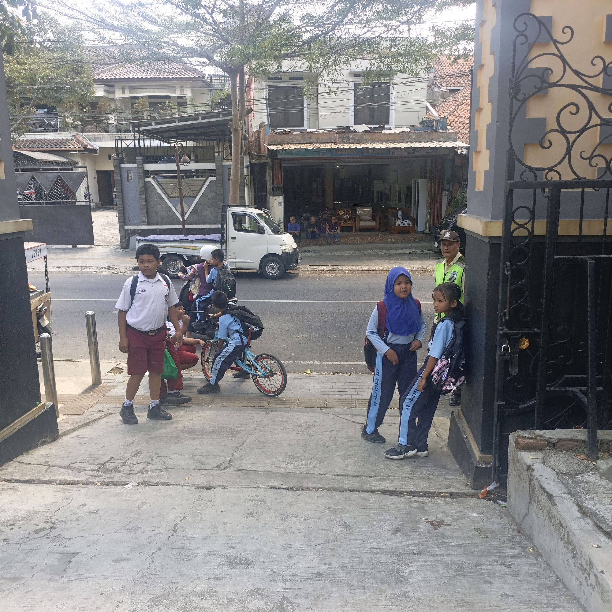 The image depicts a street scene in Salatiga, Indonesia, likely near a school. In the foreground, a young boy wearing a white collared shirt and maroon shorts stands on a sidewalk, facing left. He has a backpack on his shoulders. To his left, on the street, a white truck is parked. Behind the boy, several other children in school uniforms are visible. Two girls in blue hijabs and school uniforms are standing near a black metal gate on the right side of the image. One girl is looking at the camera, while the other is looking to her left. Behind them, a security guard in a yellow vest and a brown cap stands near the gate. Several other children are on the sidewalk and street, some near a bicycle. The environment appears to be an urban setting with buildings and trees in the background.