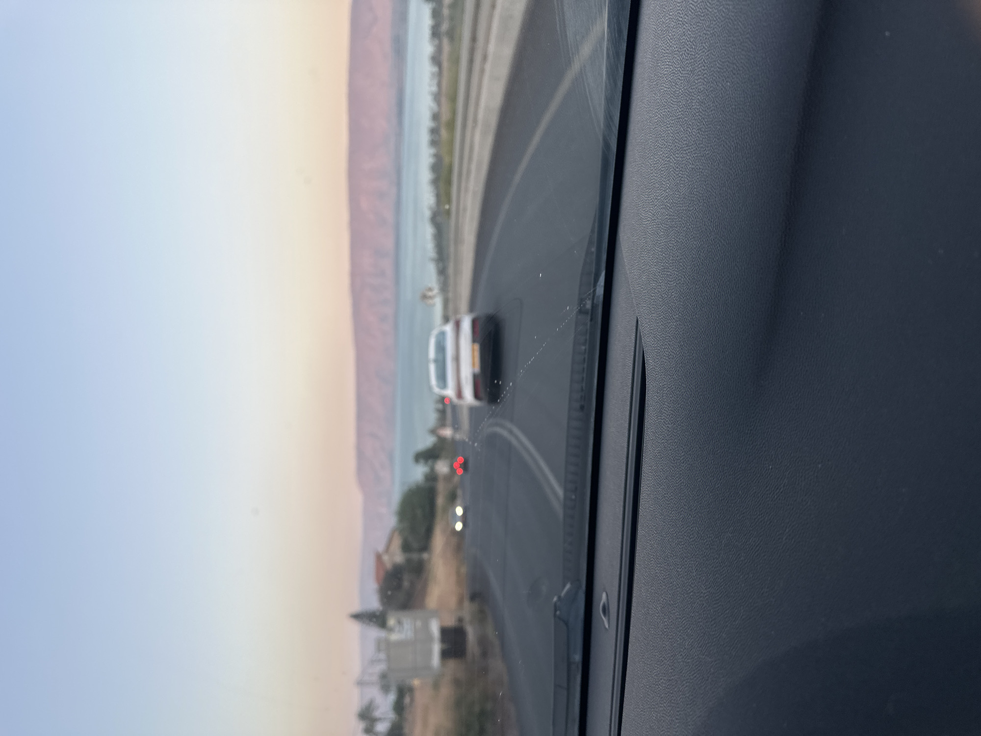 The image is taken from the interior of a vehicle looking out through the windshield. The scene depicts a road curving to the right, with a body of water and hills in the background. The sky is a gradient of pale blue to pale orange, suggesting either sunrise or sunset.
On the road, there are several cars. The closest is a white sedan in the right lane, its rear facing away from the camera. Further ahead, in the left lane, are two cars with their headlights on, one with bright white headlights and another with red taillights. The road appears to be a two-lane highway with a breakdown lane on the right.
The landscape to the left of the road shows dry, scrubby vegetation, some trees, and buildings, suggesting a rural or semi-urban setting within Emek HaYarden Regional Council, Israel. The background is dominated by a wide expanse of water, likely the Sea of Galilee, with hilly terrain on the opposite shore, colored in soft hues of pink and purple. The overall impression is a calm, scenic view during a transitional time of day.