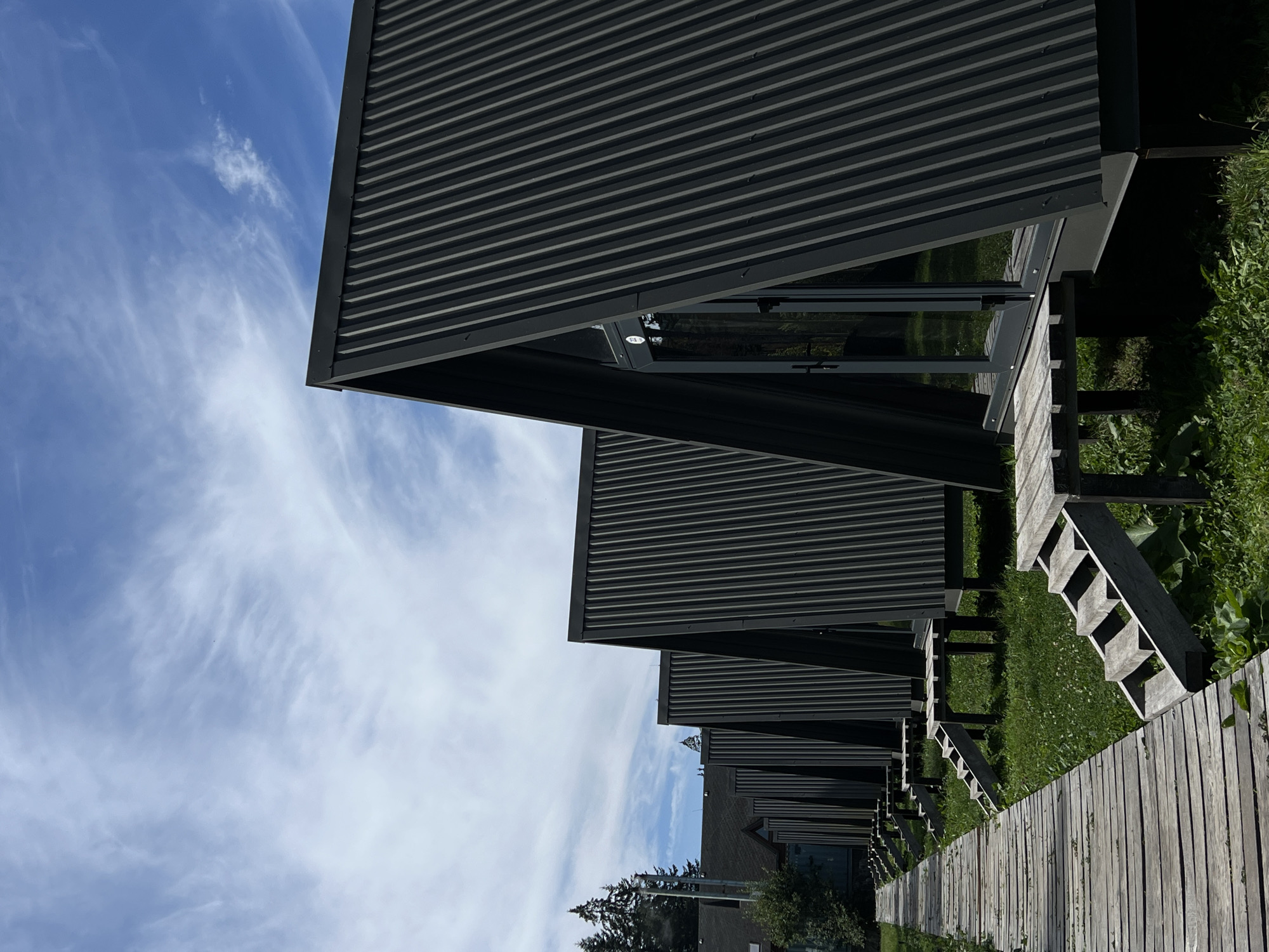 In Vorokhta, Ukraine, a row of modern A-frame cabins with dark corrugated metal roofs stands under a bright blue sky with scattered white clouds. A weathered wooden boardwalk runs alongside the cabins, leading the viewer's eye toward the structures. Each cabin features a glass door and a small wooden deck with a set of wooden steps. The cabins are set on a grassy area with some low-lying vegetation. In the background, a few evergreen trees are visible, along with a larger building with a dark roof. The scene appears to be a tranquil resort or glamping site.