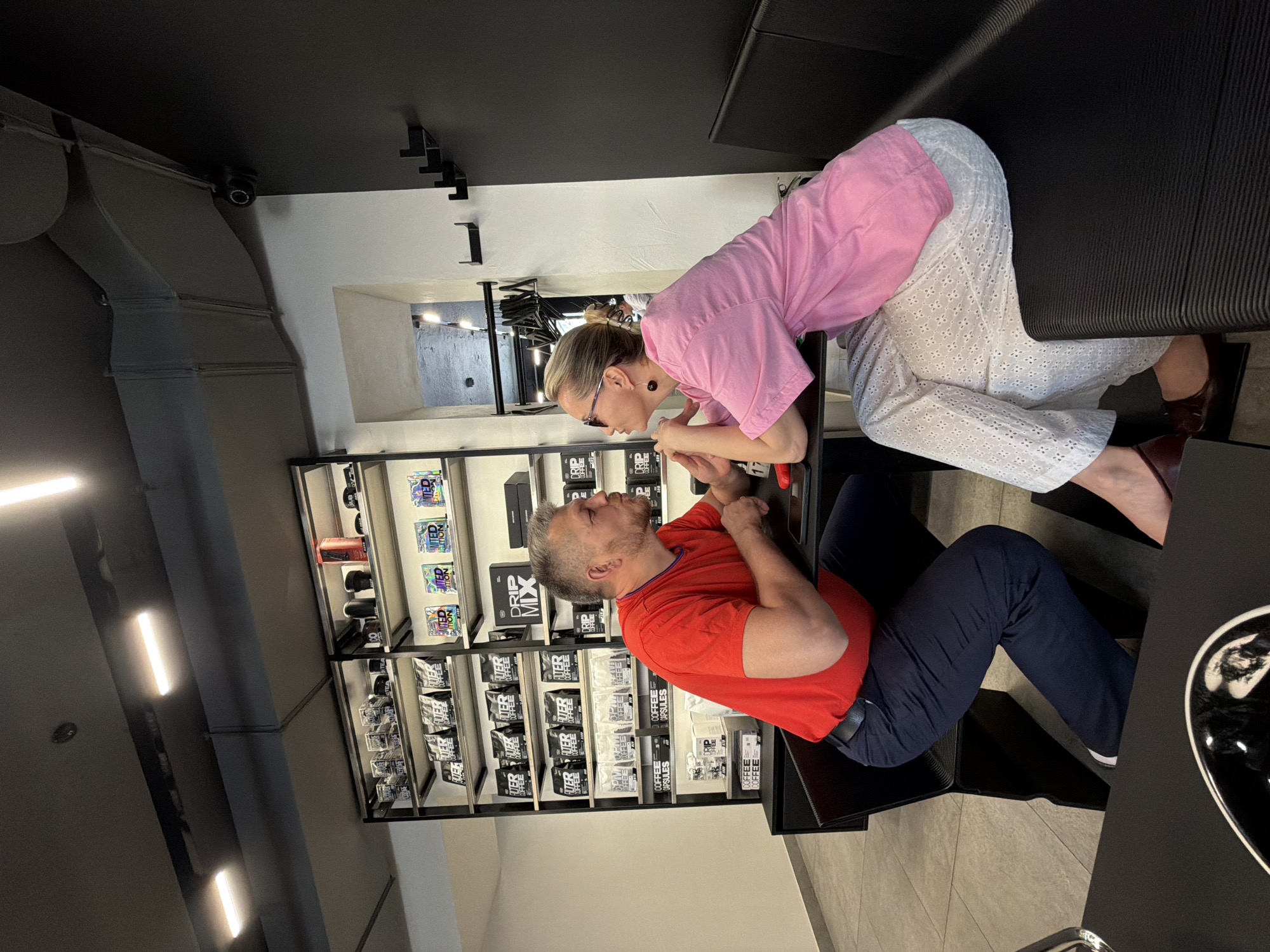 The photo shows a woman and a man at a coffee shop in Izhevsk, Russia. The woman is leaning over a table with her hands on her face, looking at the man sitting across from her. She is wearing a pink shirt and white patterned pants. The man is wearing a red shirt and blue pants. The coffee shop has shelves filled with coffee products. The interior has a modern design with black and white accents.