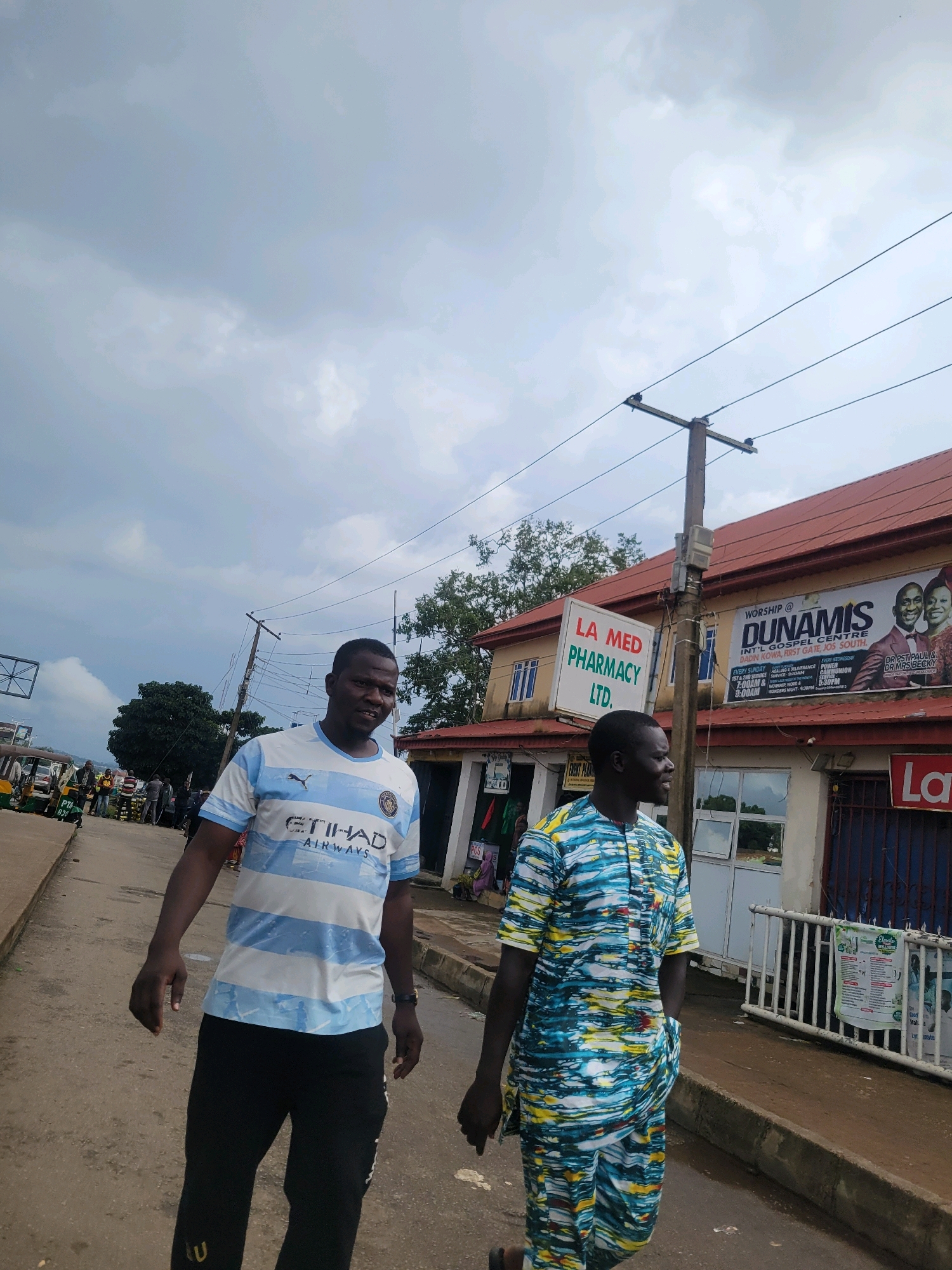 Two men are walking on a dusty road in the city of Jos, Nigeria, under a cloudy sky. The man on the left wears a white and light blue striped t-shirt with "ETIHAD AIRWAYS" logo and black pants. The man on the right wears a vibrantly patterned blue, yellow, and white African print tunic and pants. In the background, a building with a red roof has a sign for "LA MED PHARMACY LTD." and another large banner for "DUNAMIS INT'L GOSPEL CENTRE." A utility pole with electrical wires stands in front of the building. Other people and vehicles, including a three-wheeled auto-rickshaw, are visible further down the road, suggesting a commercial area.