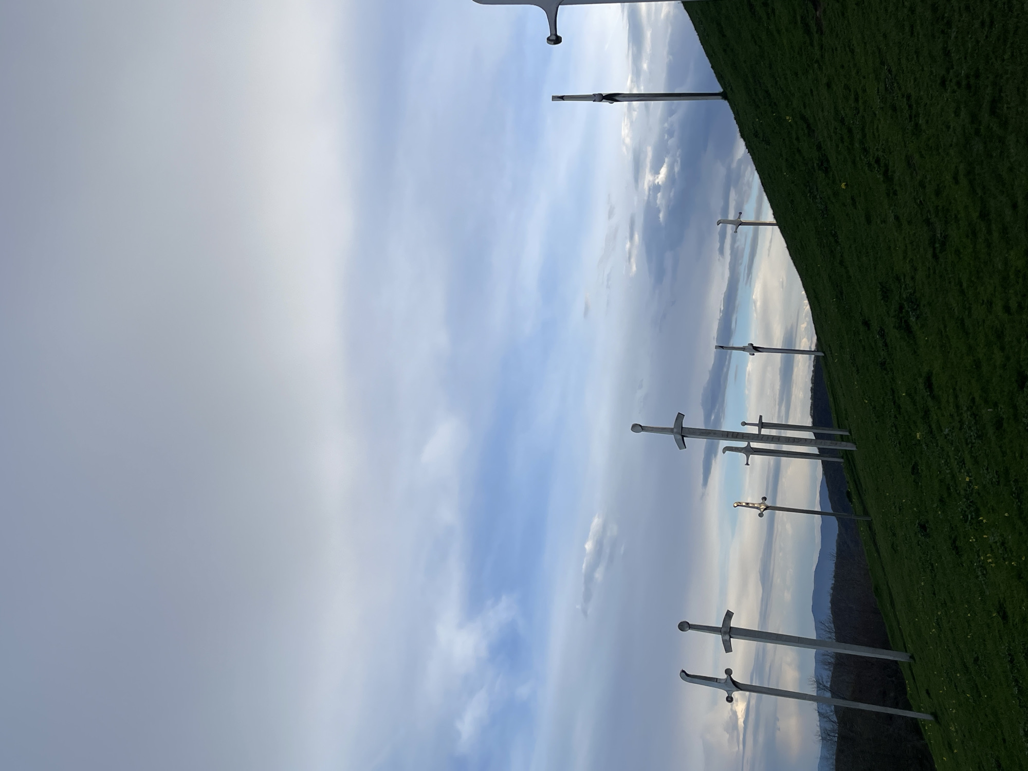The image shows a monument of several swords planted into a grassy hillside. The background consists of a cloudy sky and a distant treeline. The location is likely Zemo Nichbisi, Georgia. There are no people in the shot.