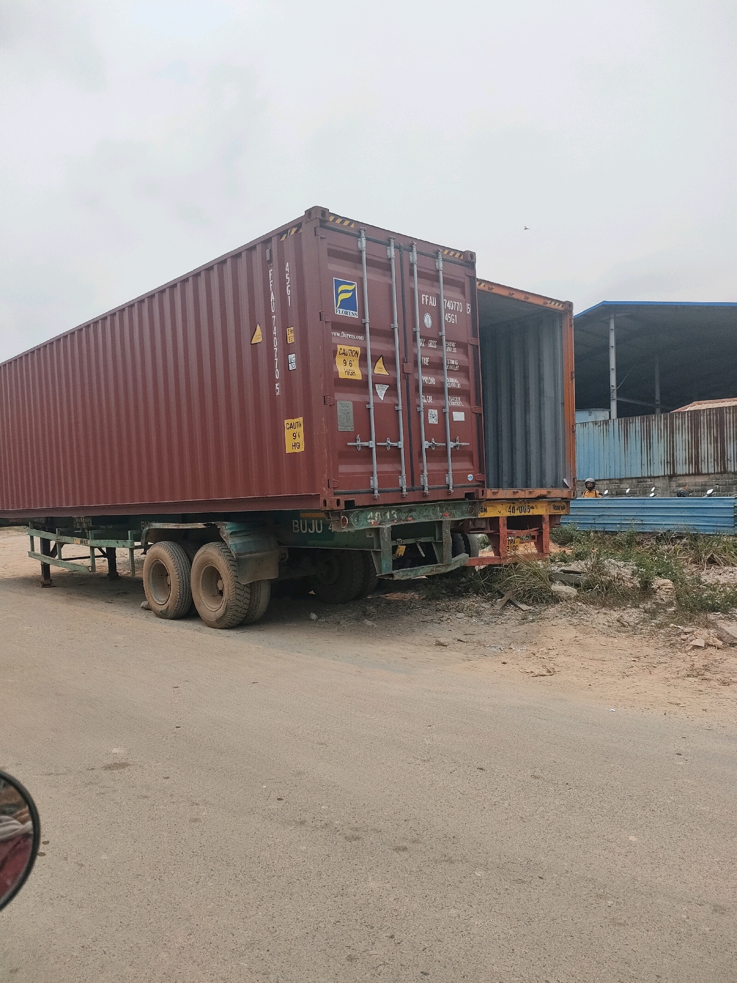 The image shows a large, dark red shipping container loaded onto a trailer with dual wheels. The trailer is parked on a dirt road, with a building and some blue metal sheeting in the background. The shipping container has markings on its side, including "F.FAU 740770 5 4561" and "FLORENS". There is an open section of the container, revealing its interior. A person in an orange shirt is visible near the blue metal sheeting in the background. The sky is overcast. The scene appears to be an industrial or logistics area in Batam, Indonesia.