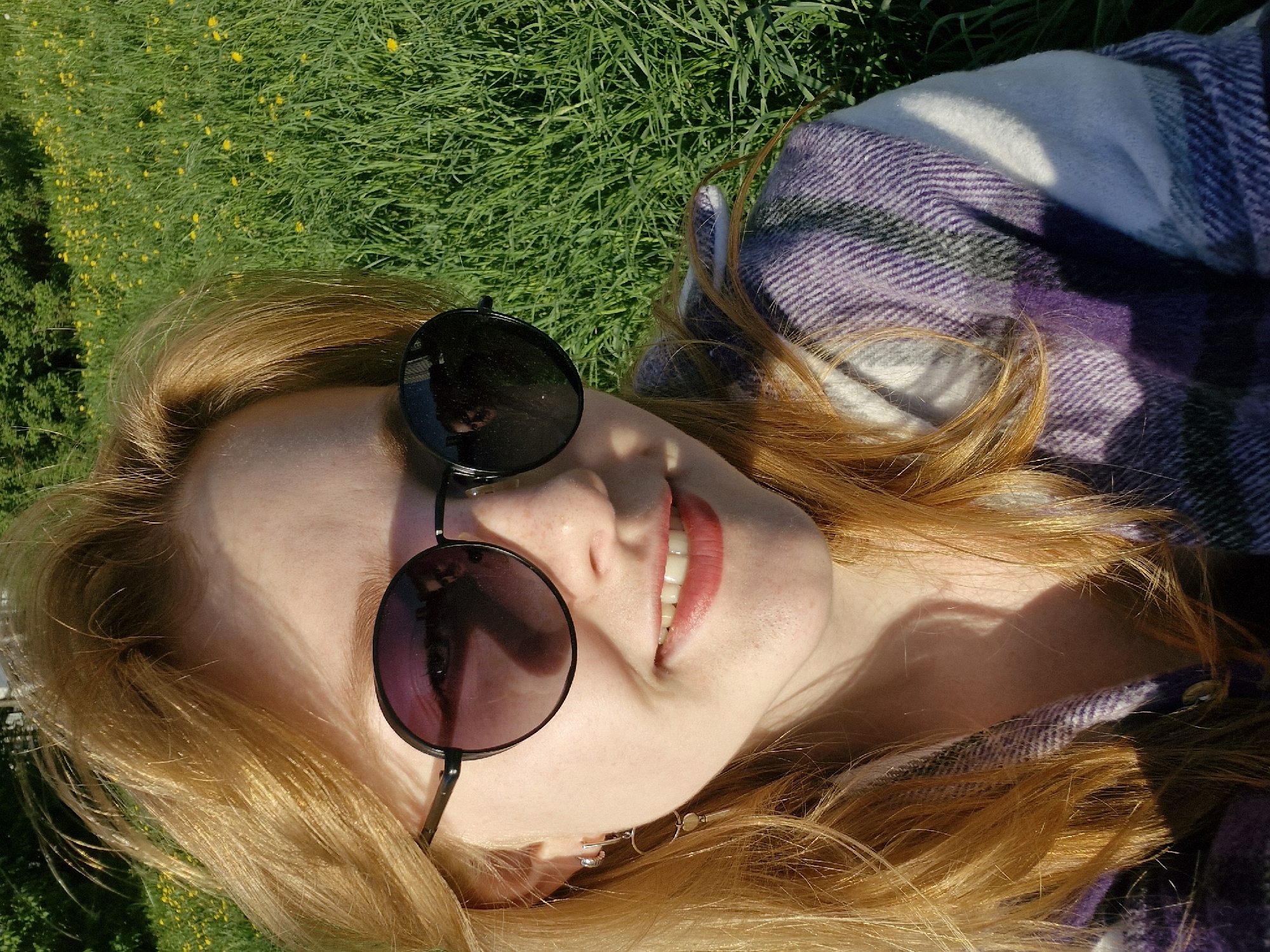A woman with red hair is lying on the grass in a park-like setting. She is wearing round sunglasses and a purple and white plaid blanket. The location is not identifiable as Yekaterinburg, Russia based on the image content. The background shows a grassy field with small yellow flowers. The woman is smiling slightly.