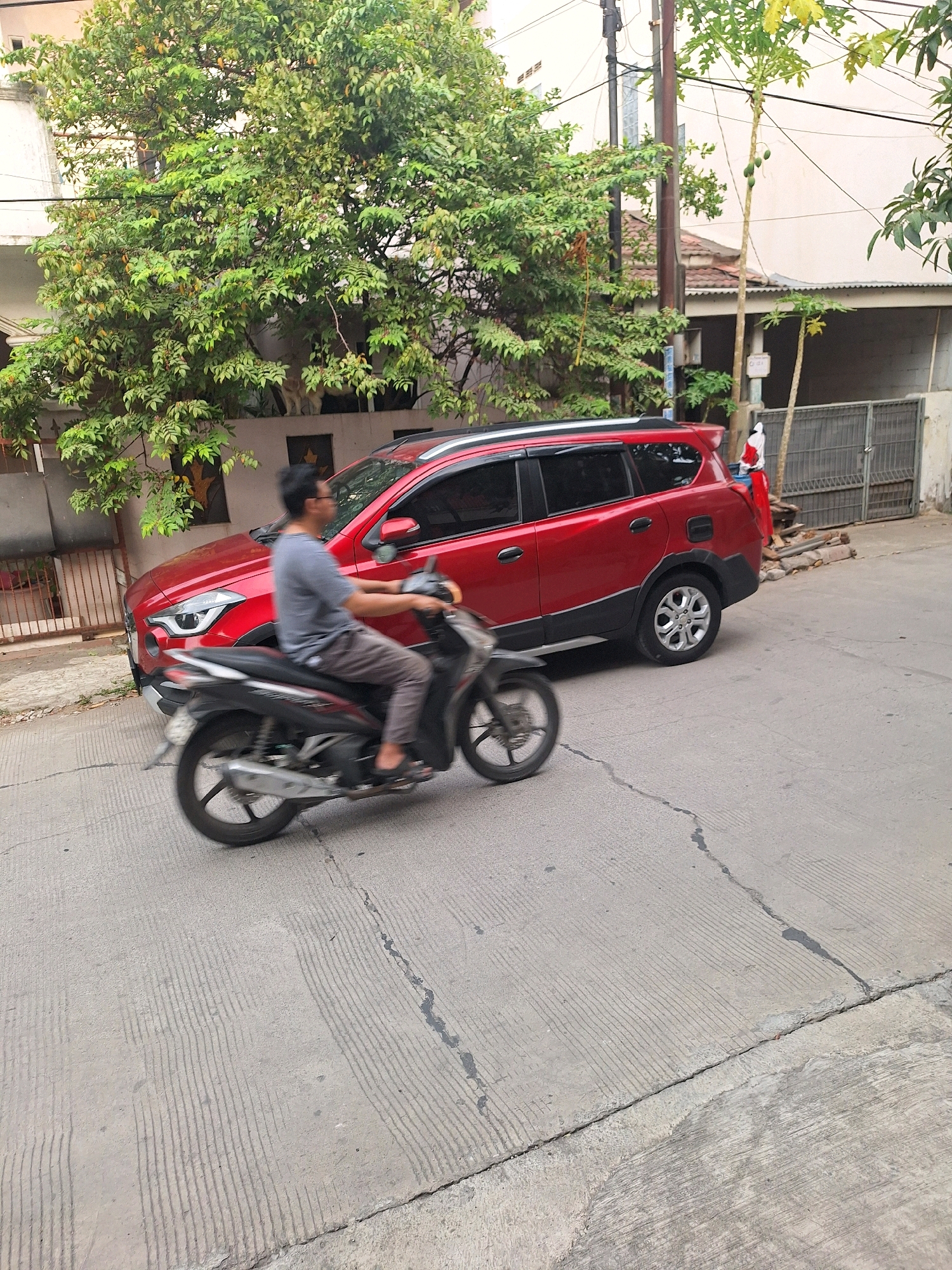In Setiamulya, Indonesia, a red crossover utility vehicle is parked on a paved street next to a building with green foliage. A man is riding a motorcycle in motion past the parked vehicle. The street has visible cracks and grooved patterns. A fence and more buildings are visible in the background.