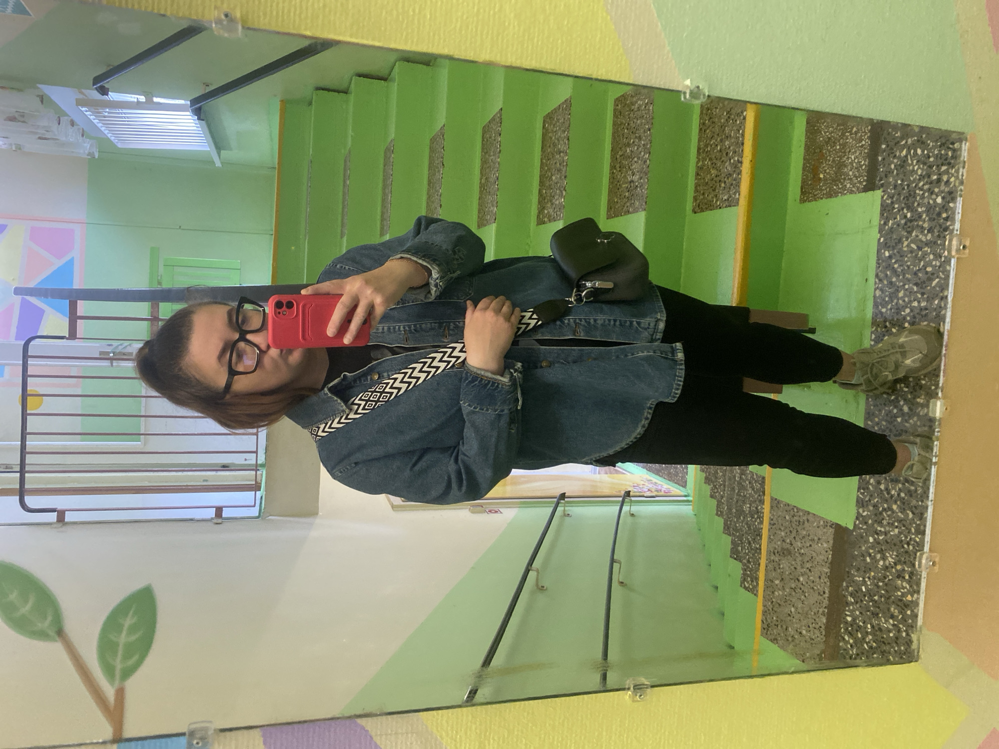 A woman is taking a selfie in a mirror at an entrance in the city of Chaikovsky, Russia. She is wearing glasses, a blue denim jacket with a black and white strap, black pants, and gray sneakers. She is holding a red phone. The mirror is mounted on a wall with green, yellow, and purple colors. The reflection shows a staircase with green steps and a terrazzo landing. A drawing of a tree with green leaves is visible in the reflection, along with a barred window.