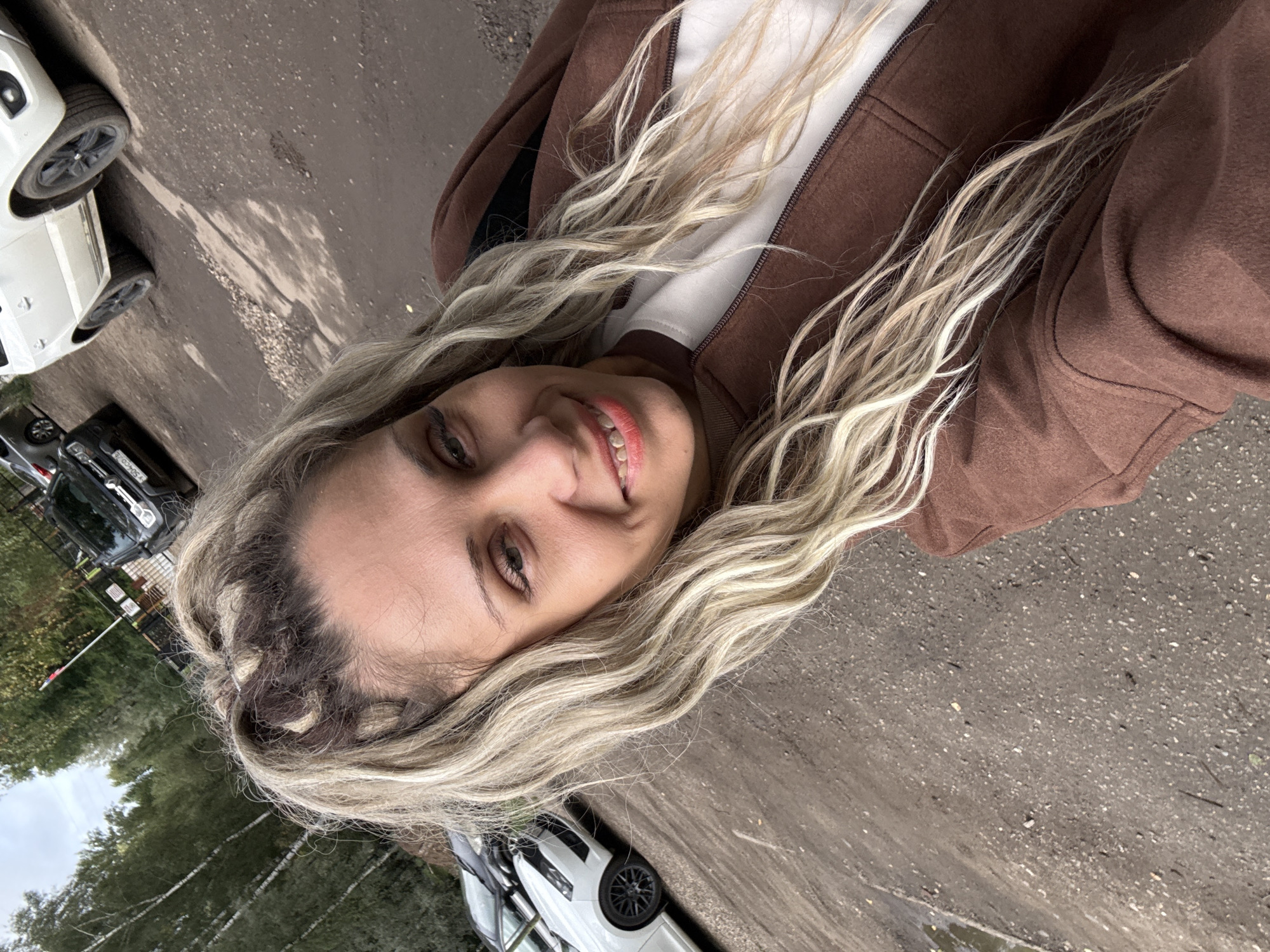 In Rusanovo, Russia, a woman with blonde, wavy hair styled in braids stands outdoors, taking a selfie. She is smiling and looking at the camera. She is wearing a brown jacket over a white shirt. Several cars are parked in the background, including a black Renault and a white SUV. The ground is dirt and gravel, with some puddles of water. Trees and a fence are visible in the background.