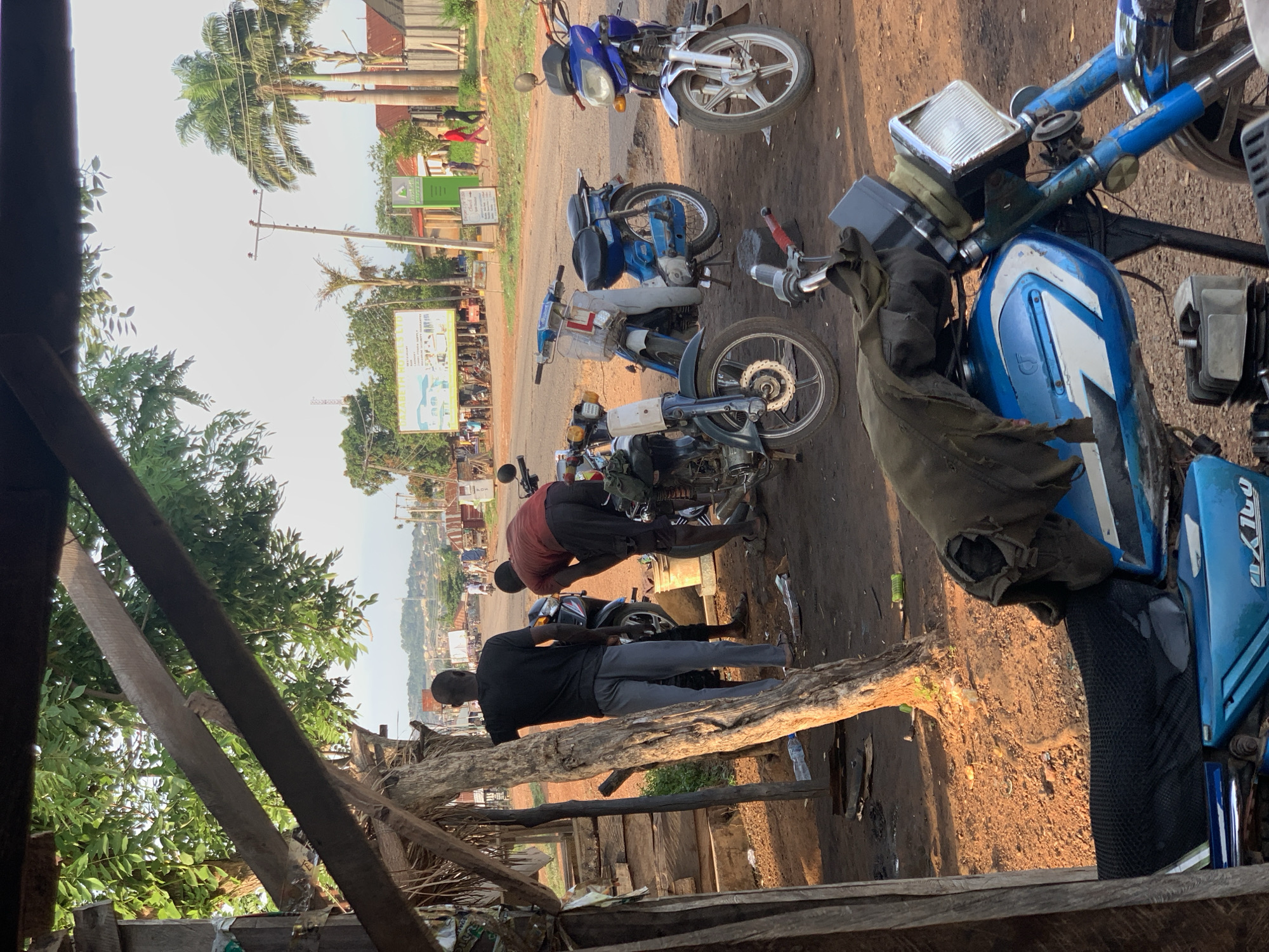 The image shows a street scene in Kabba, Nigeria. Several motorcycles are parked on a dirt surface. Two men are working on one of the motorcycles. One man is wearing a red shirt and the other is wearing a black shirt and gray pants. In the background, there are trees, signs, and buildings visible. The perspective is from within a wooden structure.