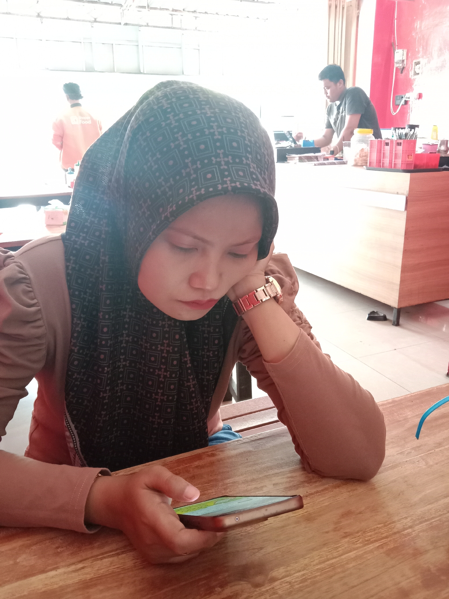 A woman wearing a hijab is seated at a wooden table in Kaliwungu, Indonesia. She is looking down at a smartphone in her hand, with her head resting on her other hand. She is wearing a brown top and a watch. In the background, another person in an orange shirt is visible near a window, and a man is working behind a counter. The setting appears to be a restaurant or cafe.
