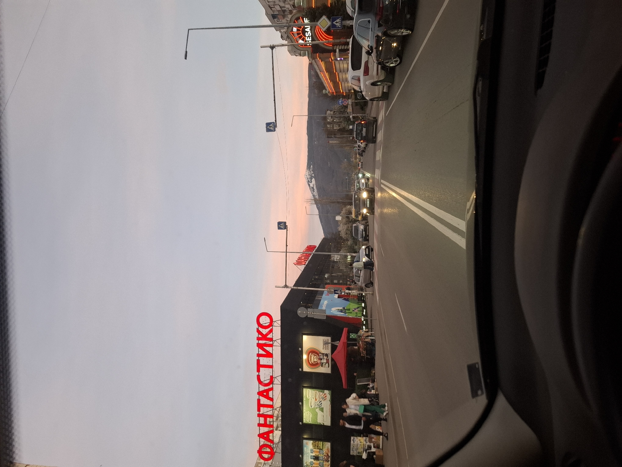 The image shows a view from inside a car, looking out onto a street in Sofia, Bulgaria. The most prominent feature is a large "ФАНТАСТИКО" (Fantastico) supermarket sign in red Cyrillic letters. The supermarket building is visible behind the sign, featuring advertisements and images. The street has multiple lanes of traffic with cars parked along the side. There are pedestrians walking near the supermarket. Further down the street, there are other buildings and businesses, including one with a neon sign that says "KESAFE." In the background, there is a view of mountains. The sky is light pink and gray, suggesting it is either dawn or dusk. There are streetlights and pedestrian crossing signs along the road. The interior of the car is visible on the right side of the image.