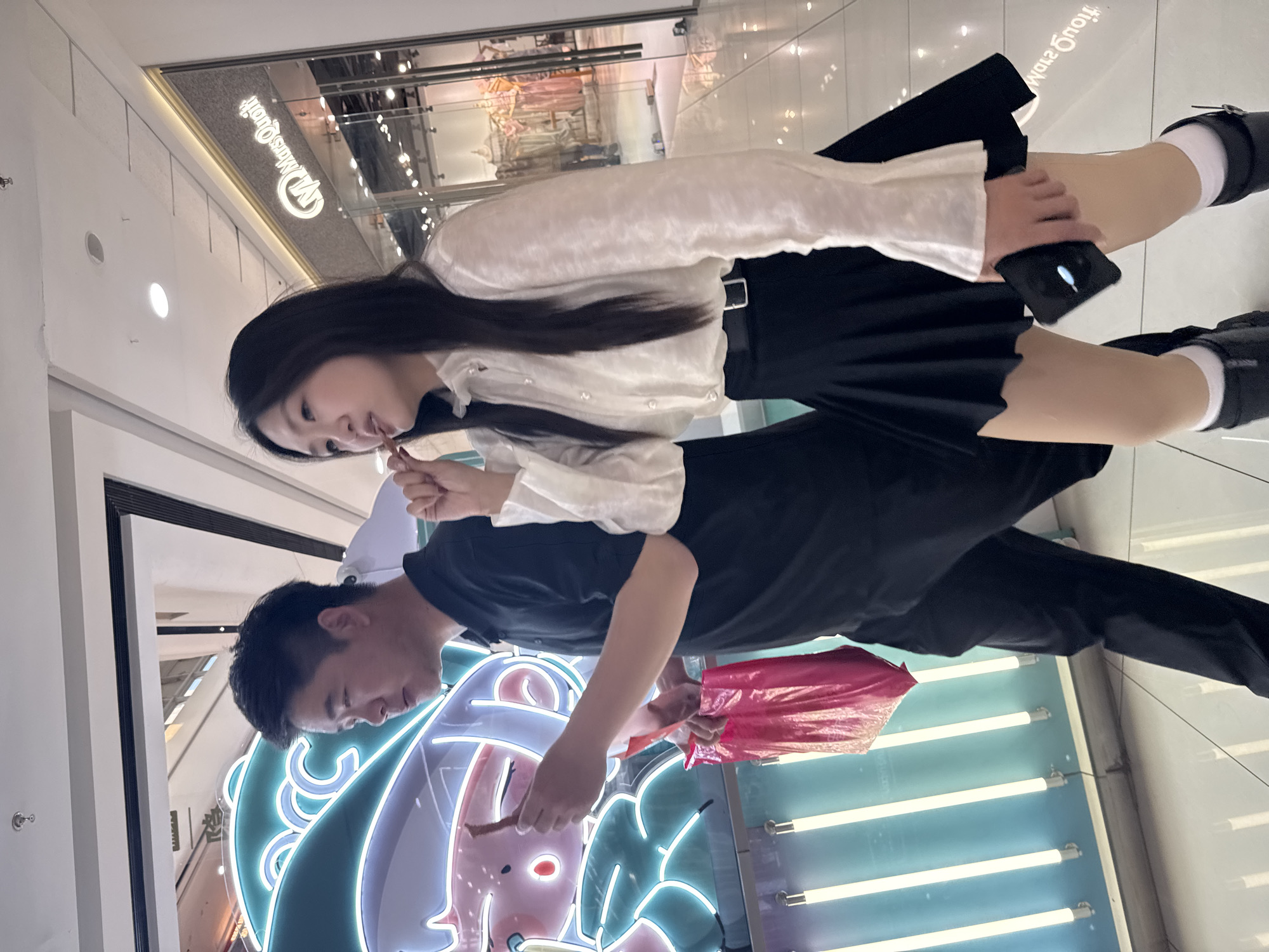 Scene in Taihe District, China, showing a young couple interacting with a cartoon advertisement within a shopping mall. The woman, dressed in a white blouse and black skirt, is eating something with a stick. She holds a phone in her other hand. Next to her, the man in a black shirt and pants is holding what appears to be a pink shopping bag. Both individuals are looking towards a brightly lit, cartoon-themed display with neon accents. Behind them is a store with “Mrs Quitt” in English letters. The floor has shiny tiles.