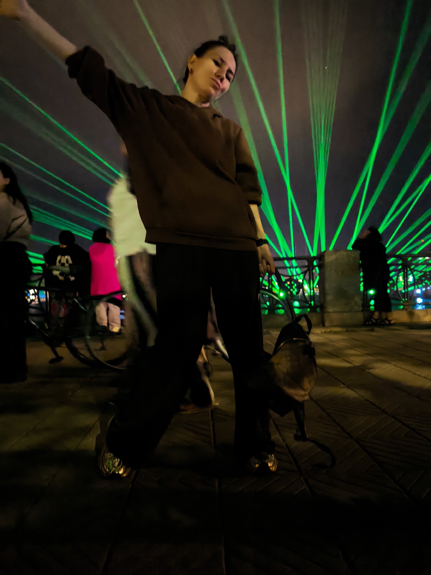 A wide-angle, low-angle shot captures a night scene with a person in the foreground, arms outstretched and head tilted back, illuminated by a display of green laser lights. The person, with dark hair pulled back, wears a brown hooded sweatshirt and dark pants. Their right arm is raised high. To the left of the foreground figure, a second person stands blurred, partially visible. Further in the background, a group of individuals and a bicycle are present. One person in the background wears a bright pink jacket. The environment appears to be an outdoor urban setting with paved ground and a metal railing. The green laser lights project upwards and outwards, creating a dramatic visual effect against a dark sky. The scene is likely located in Riga, Latvia, near a waterfront area, suggested by the faint reflections of lights in the background. The overall atmosphere is dynamic and possibly festive, given the laser light show.