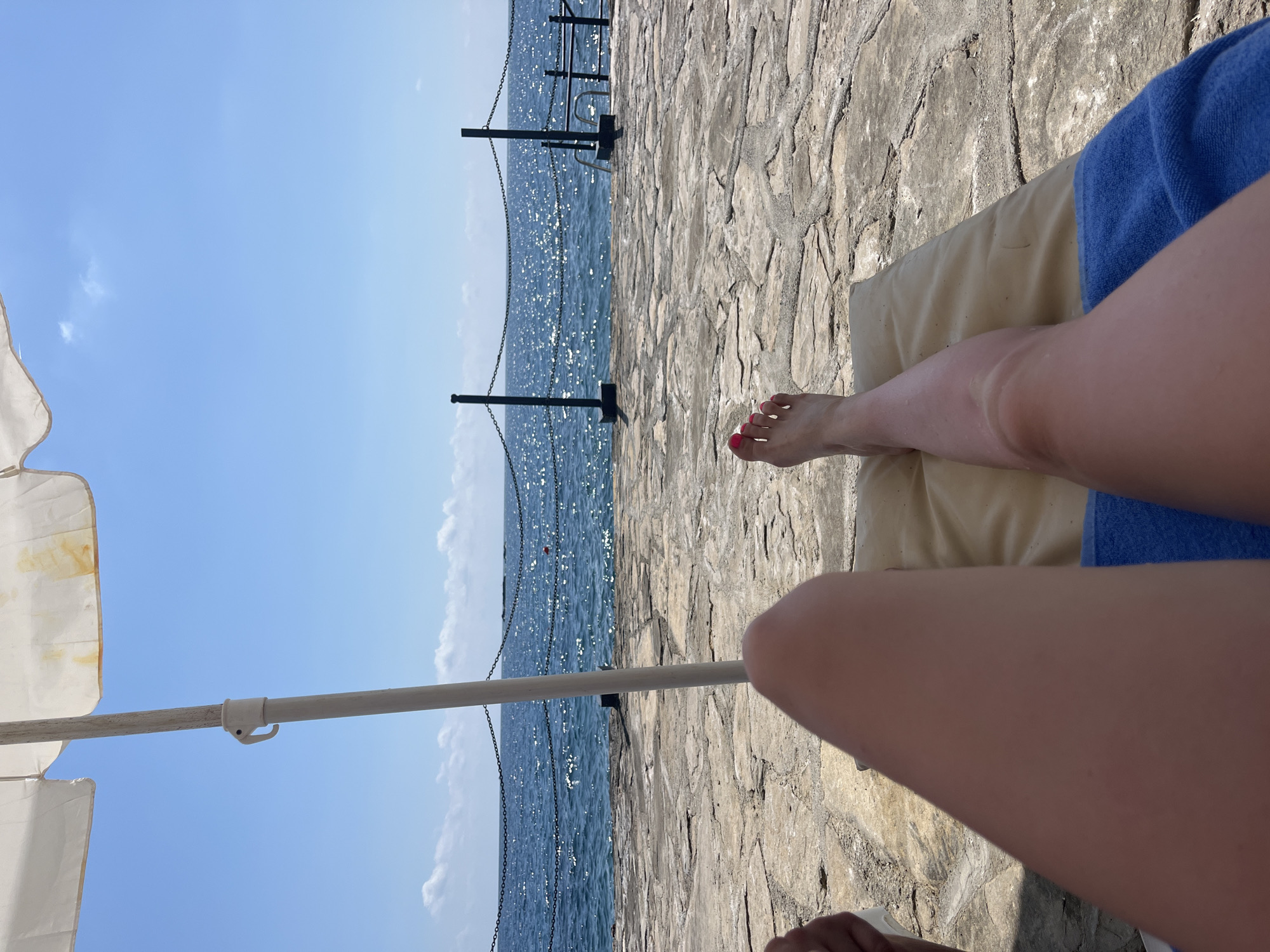 A first-person perspective shot from a lounge chair shows a person's legs and feet with bright pink toenails. They are lying on a beige cushioned lounge chair with a blue towel underneath. The person is on a stone patio overlooking the blue ocean. A white beach umbrella provides shade. In the background, a metal railing with a chain is visible, separating the patio from the water. Further out, there are a few small white clouds in the bright blue sky. This scene is consistent with a beach or poolside area in Alanya, Turkey, a popular tourist destination known for its Mediterranean coastline and sunny weather.