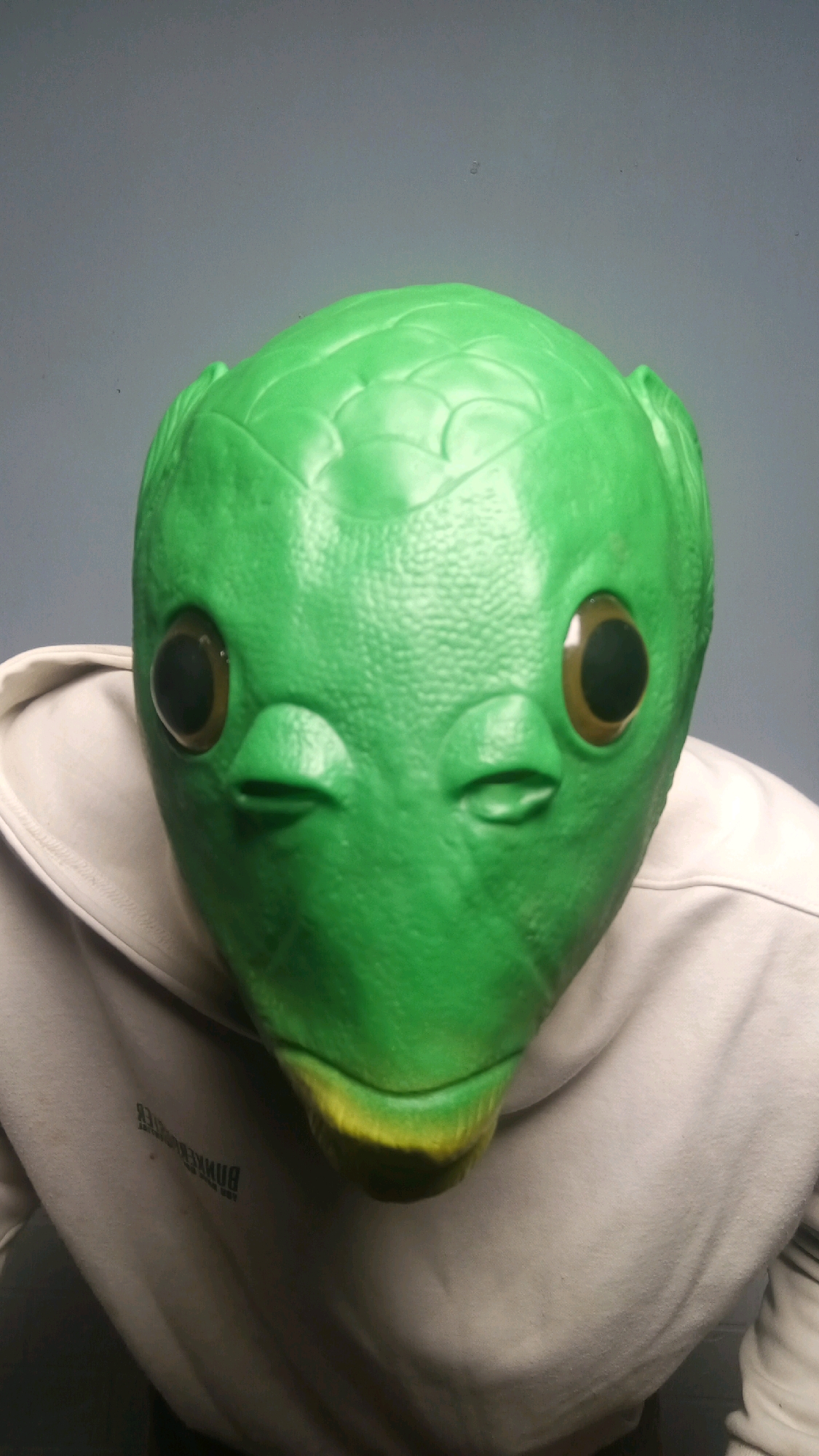 A close-up, head-on studio portrait shows a person wearing a bright green, frog-like mask. The mask has large, round, dark pupils with light brown irises, small slits for a mouth, and textured skin. The mask is pulled over a light-colored hooded sweatshirt. The background is a solid, neutral gray. The image is well-lit, with the mask as the sole focus. There is no indication of the city of Kebumen, Indonesia, or any other specific location.